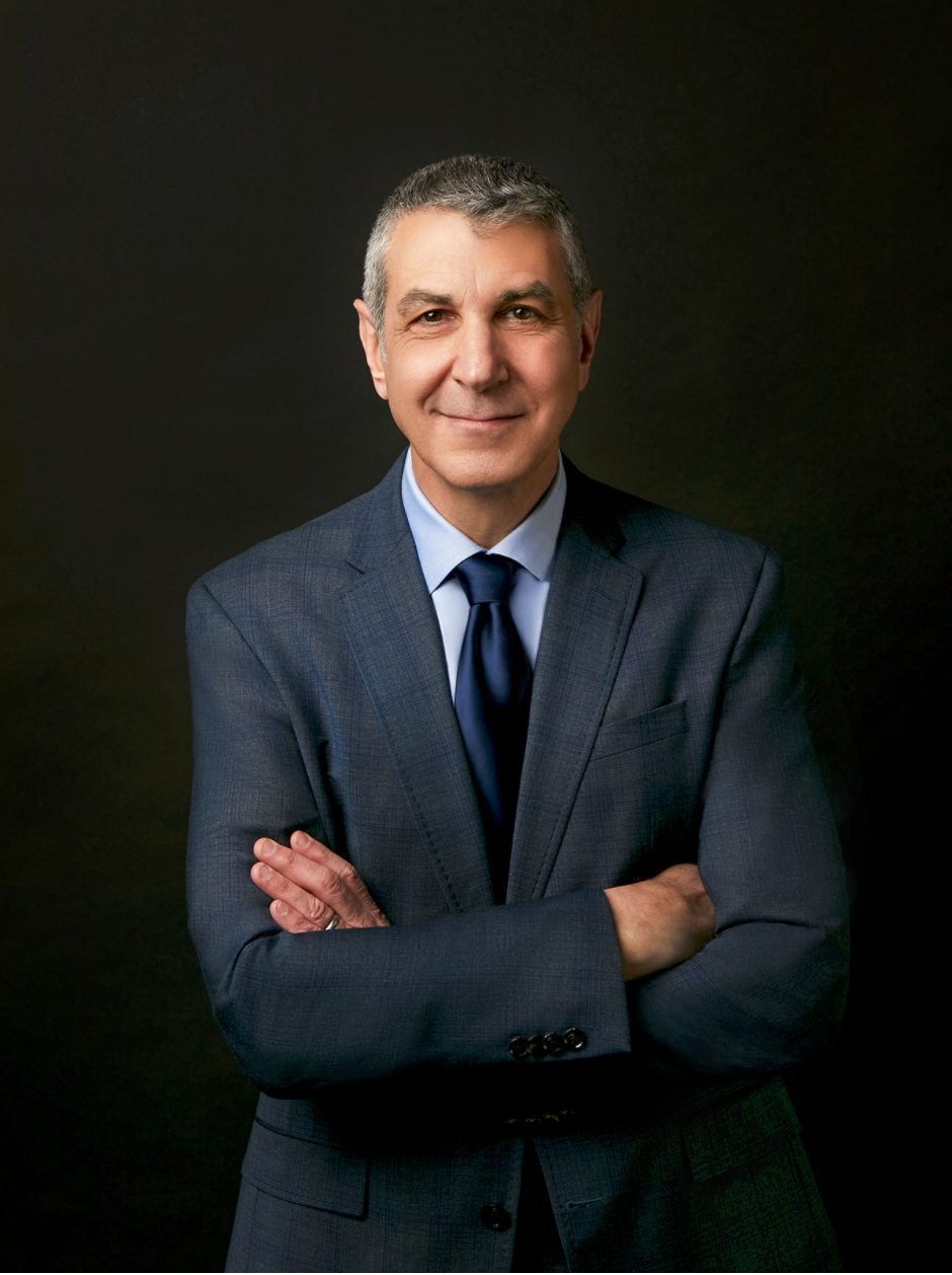 A mature male business consultant wears a blue business suit with arms crossed as he faces the camera with a soft smile for his professional headshot