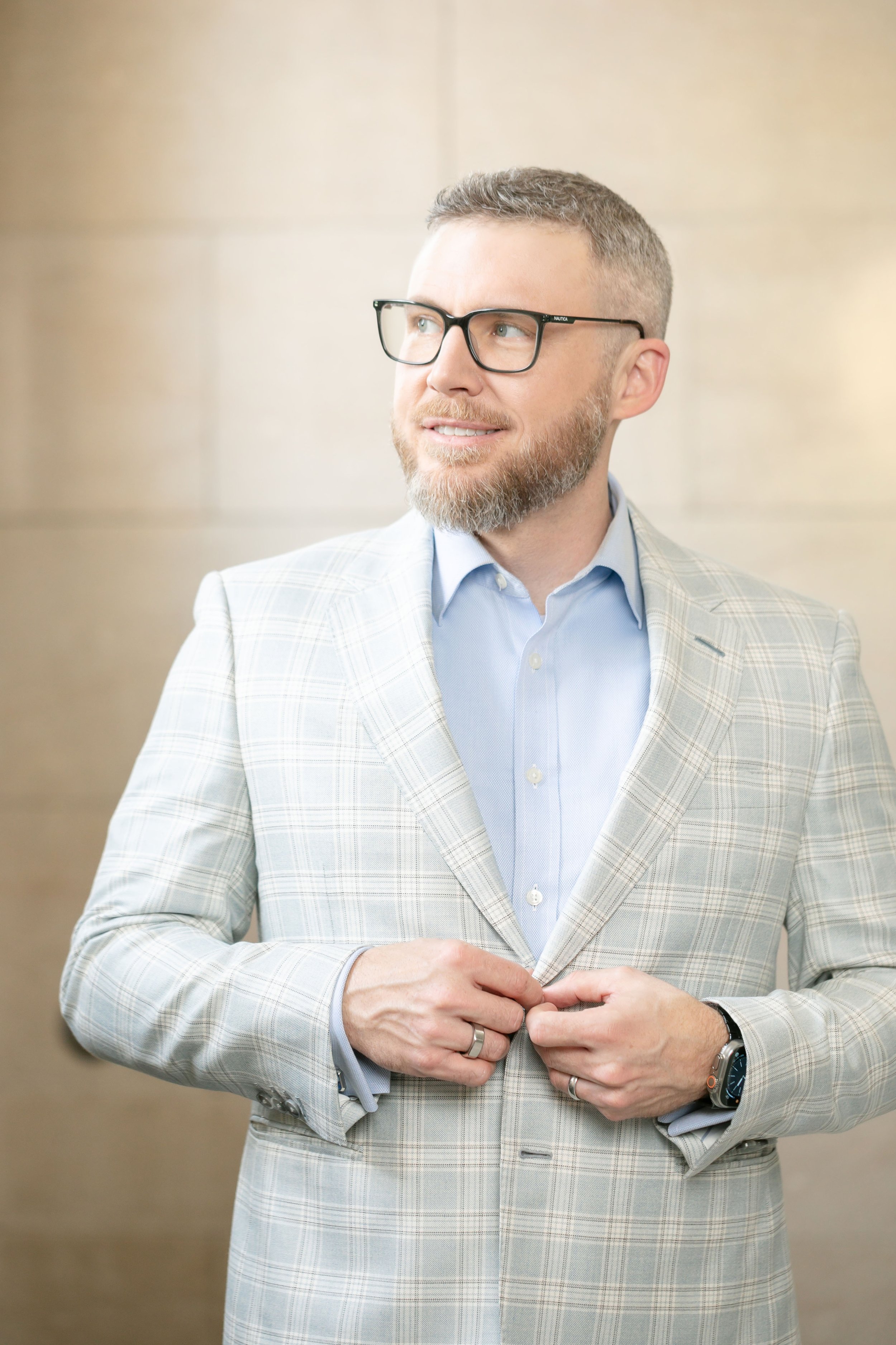 A male lawyer with glasses and a beard is adjusting his light gray plaid blazer, standing against a beige wall.
