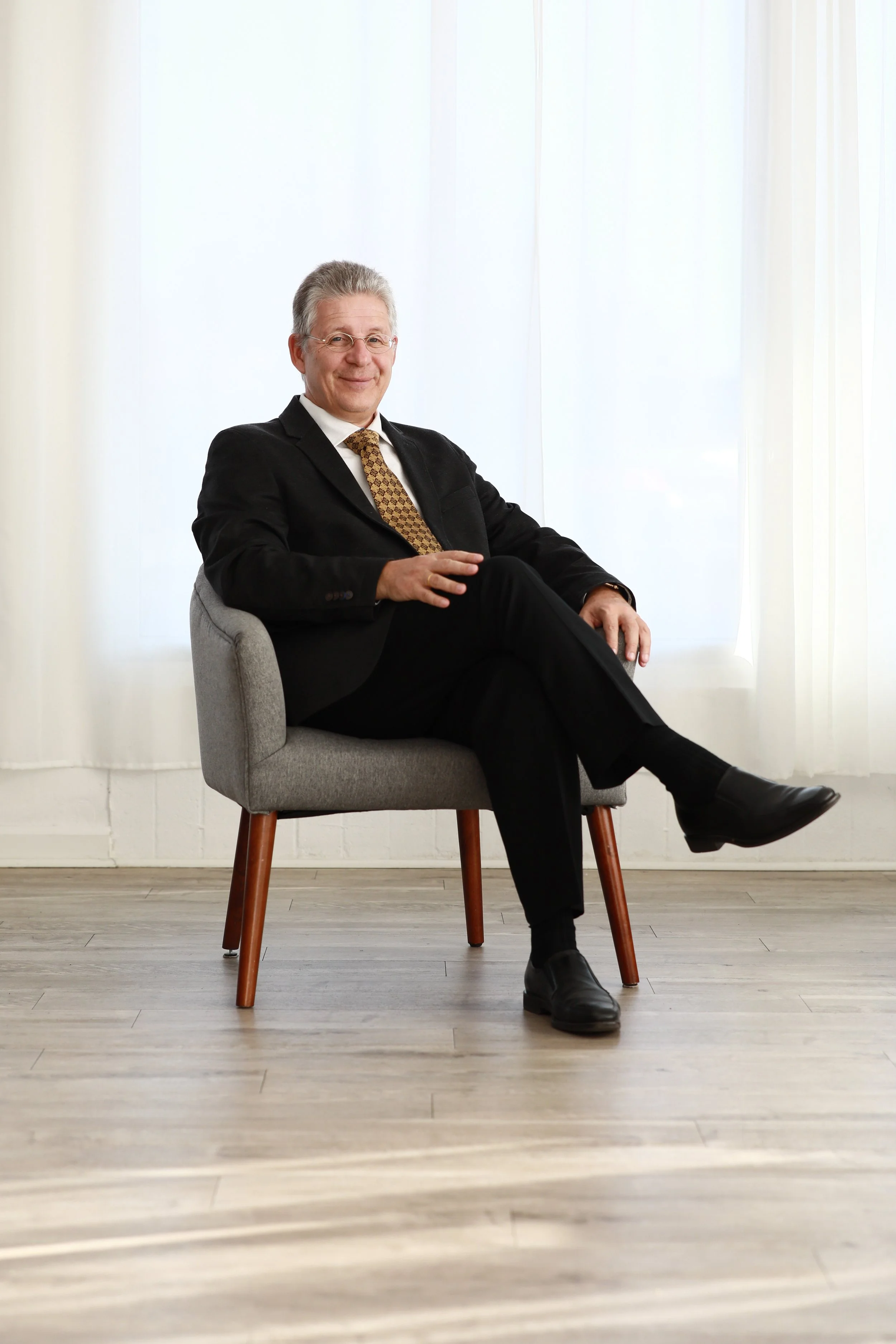 A middle-aged man dressed in a black suit and tie, sitting on a modern grey armchair with wooden legs, smiling confidently with one leg crossed over the other in a bright, minimalistic room with sheer white curtains and wooden floor for his headshot.