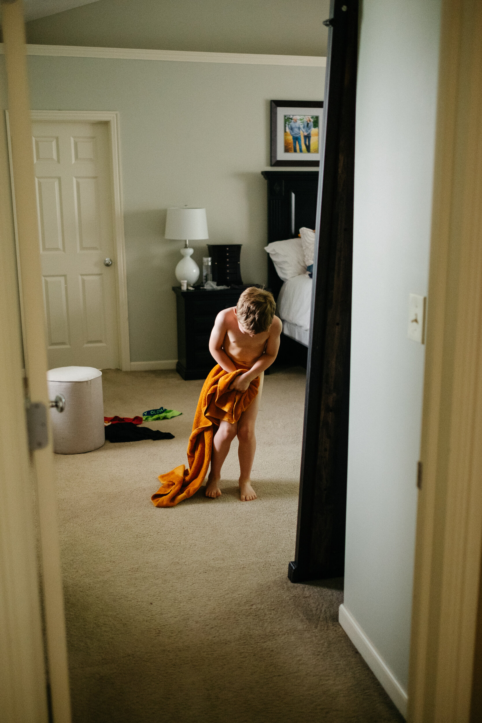 A young boy standing in a bedroom, holding an orange towel and wrapping it around himself while looking down.