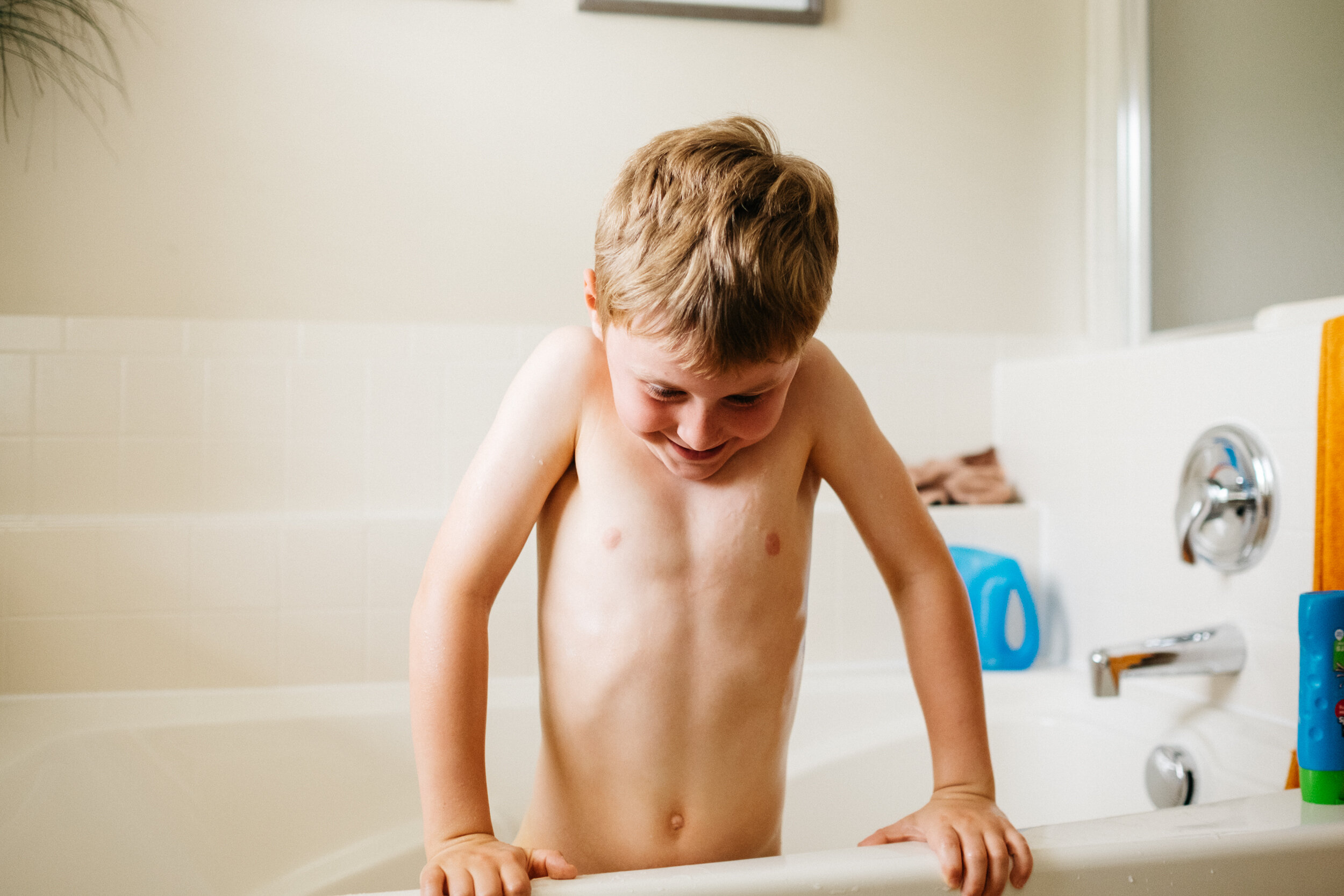 A young shirtless boy with short light brown hair taking a bath in a tub, smiling and leaning forward with his hands on the edge of the tub.