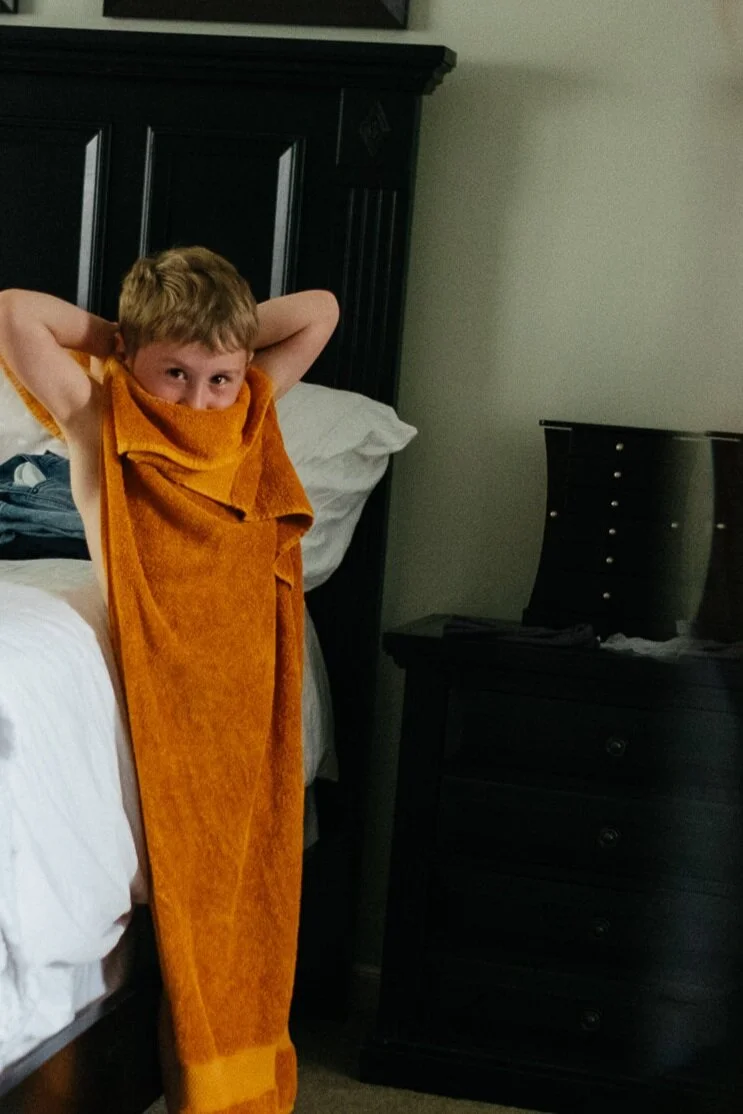 Young boy with short light brown hair standing next to a bed with white sheets, wrapped in an orange towel around his neck and shoulders, looking at the camera with hands behind his head in a bedroom with dark furniture.