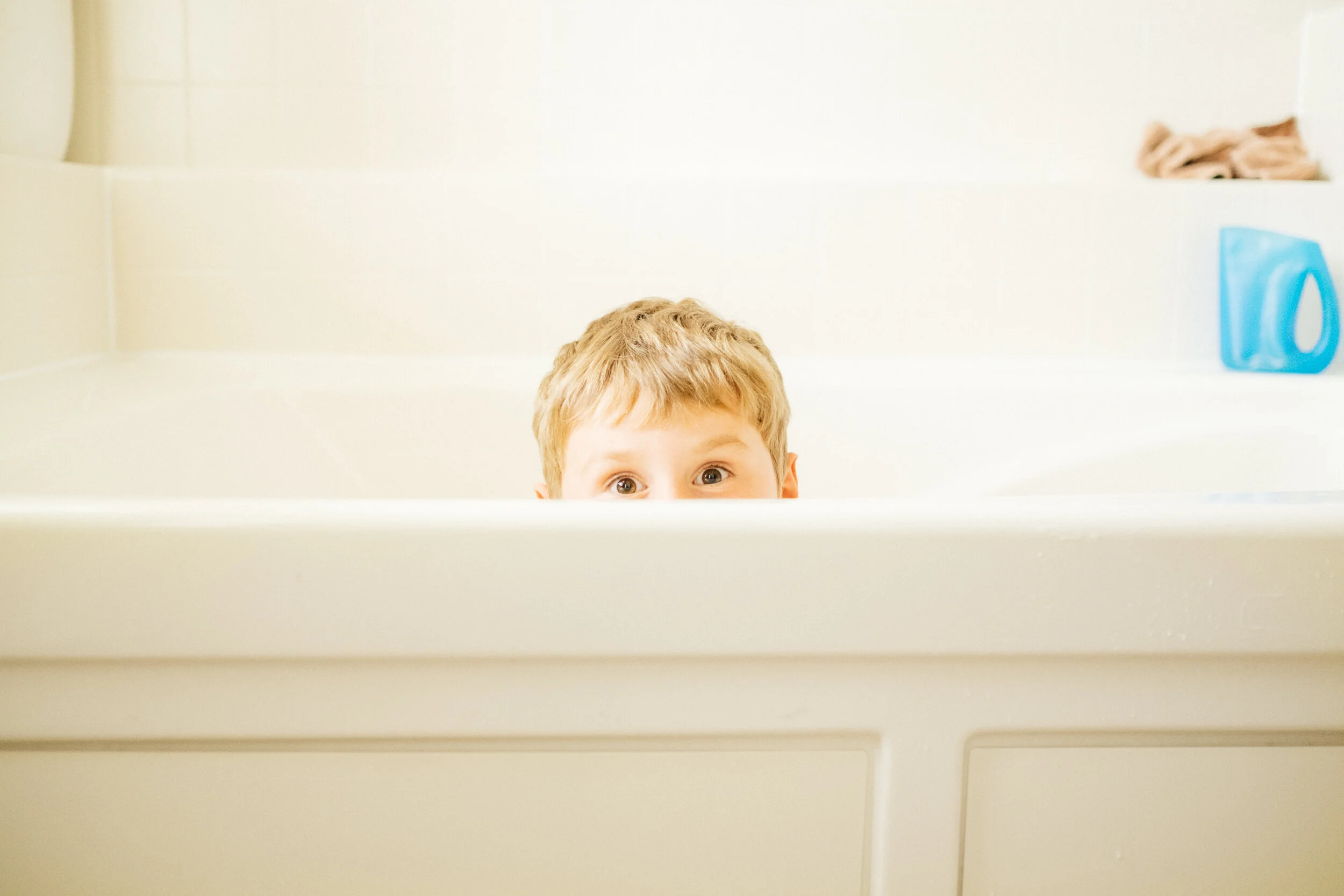 A young boy peeking over the edge of a bathtub with wide eyes, with bathroom items like a laundry detergent bottle and a towel in the background.