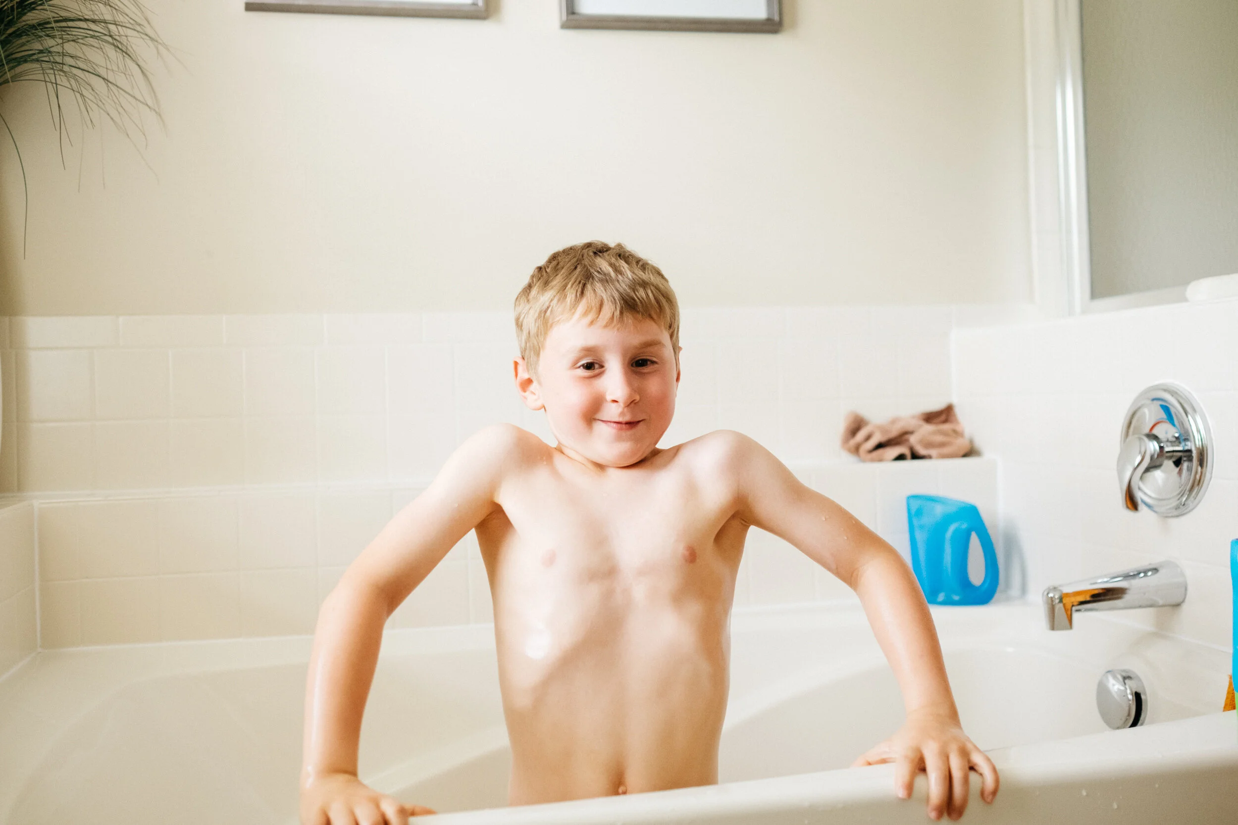 A young boy with wet hair and no shirt standing in a bathtub, smiling at the camera.