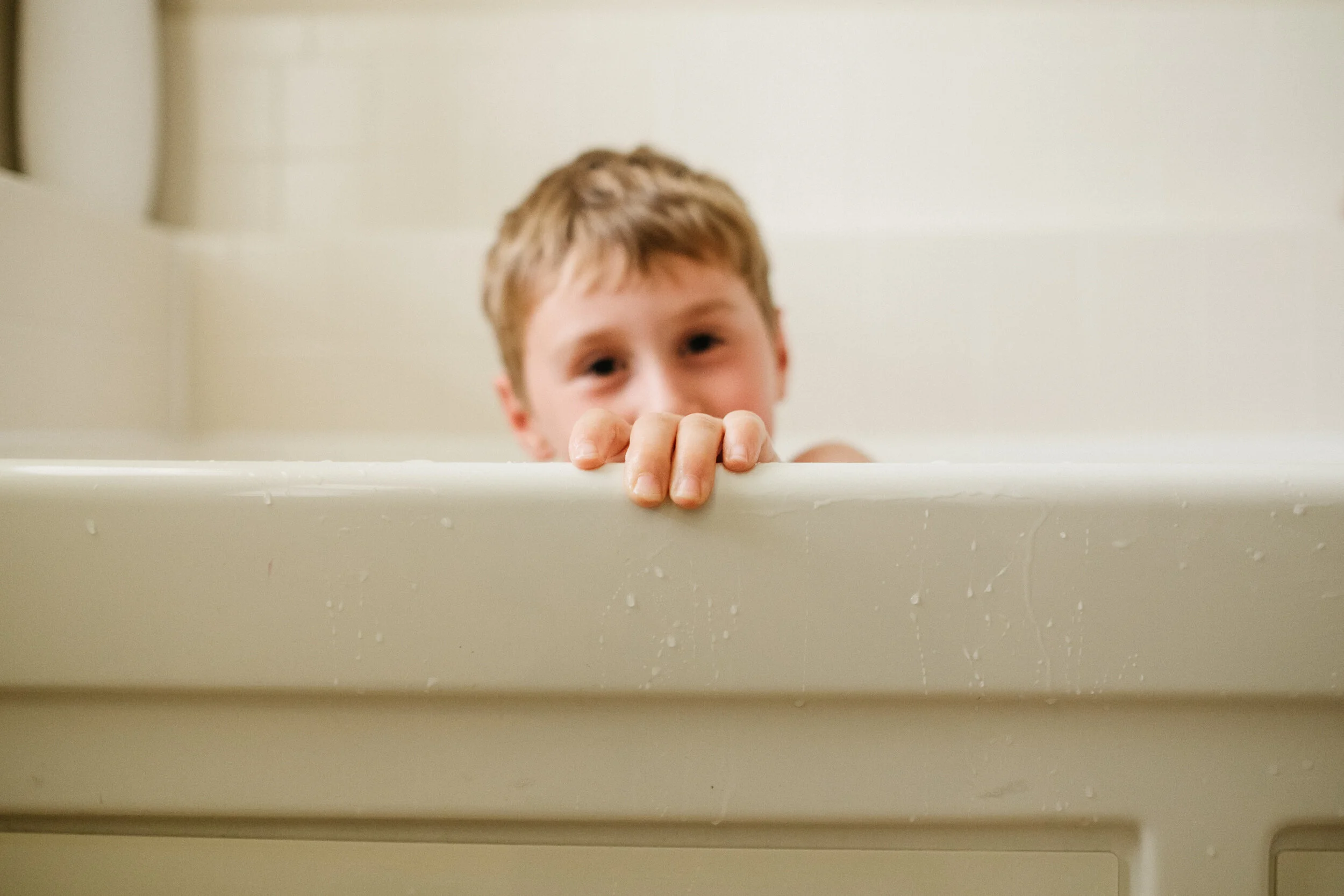 Young boy leaning over the edge of a bathtub, gripping the side with his hand, looking into the camera with a mischievous expression.