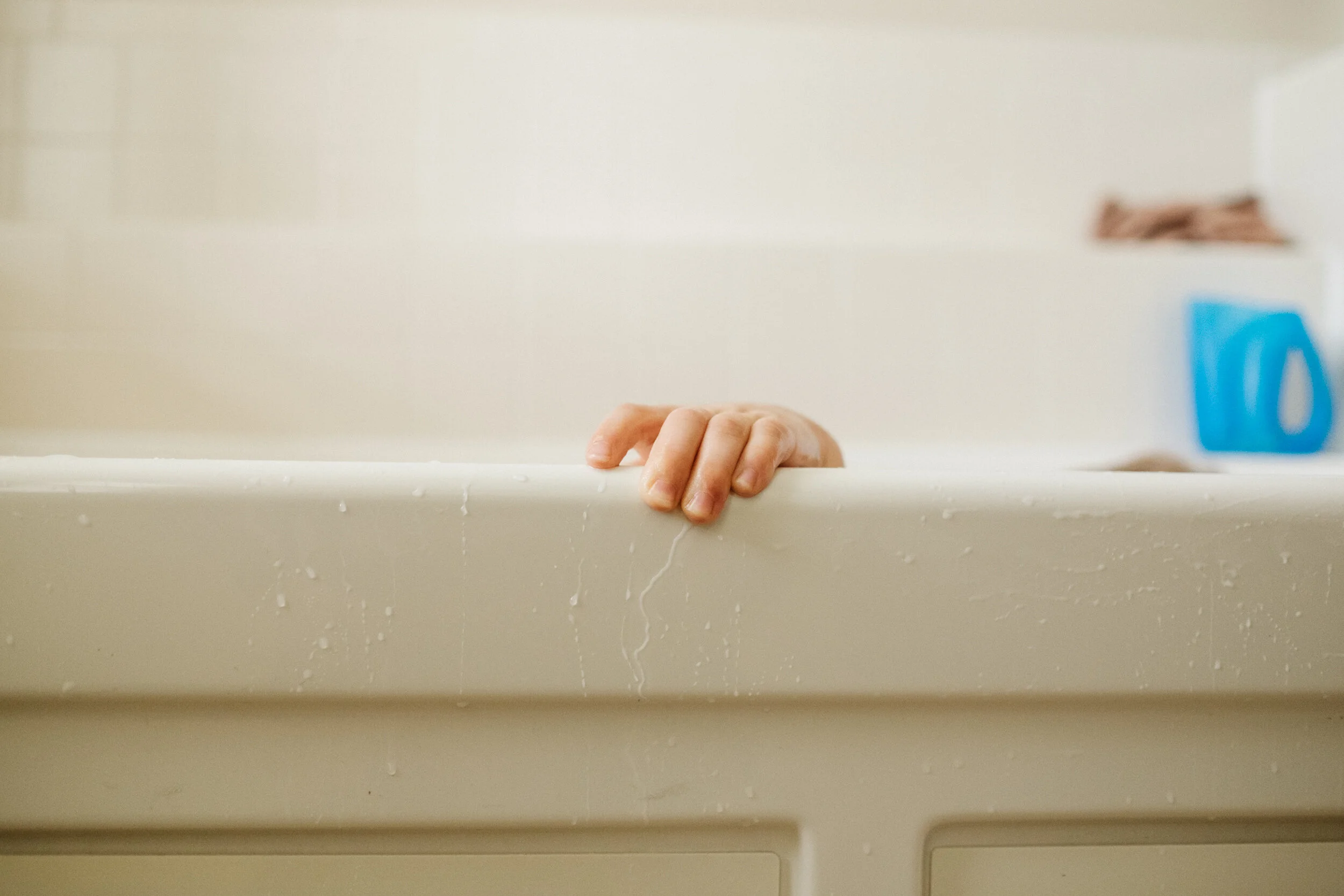 A hand peeking out from behind a bathtub edge, with water droplets visible on the surface, in a bathroom setting.