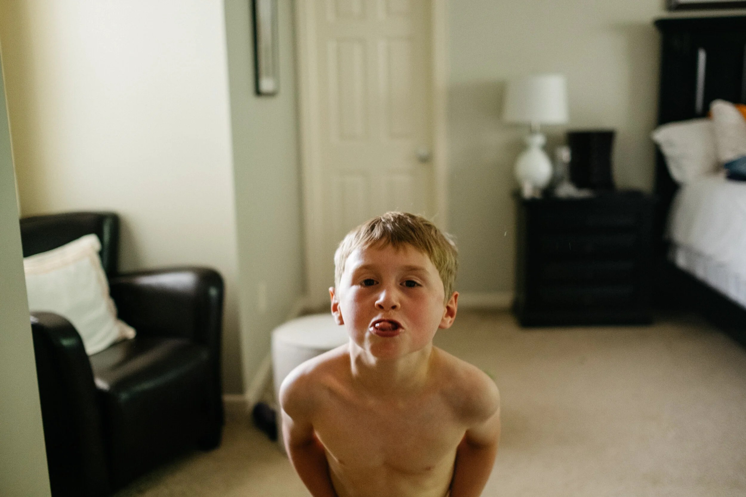 A shirtless young boy with short hair making a funny face with his tongue out in a bedroom with a bed, nightstand, and armchair.