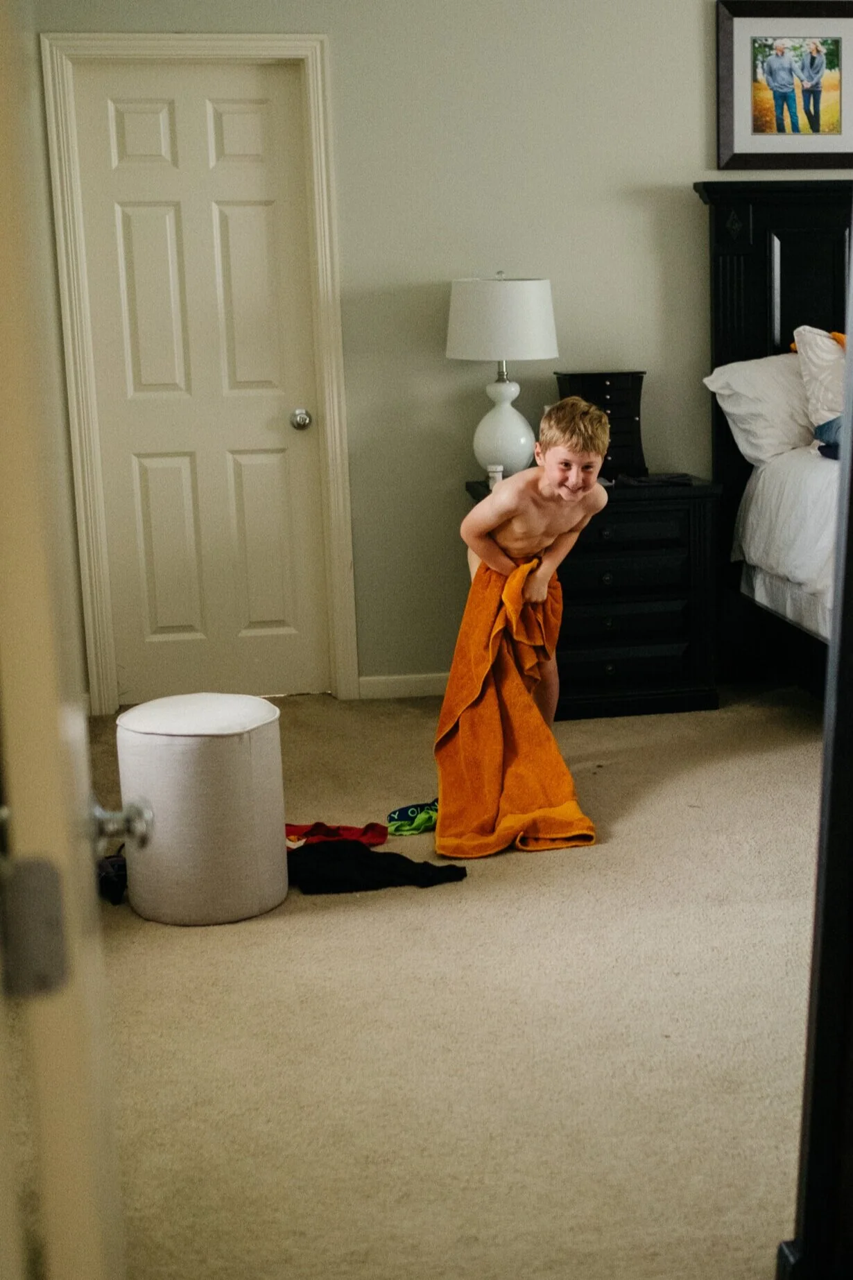 A young boy with a towel wrapped around his waist, standing in a bedroom, smiling and leaning forward. There is a white ottoman, a pair of socks, and some clothes on the floor nearby. The room contains a bed, a black dresser, a nightstand with a lamp