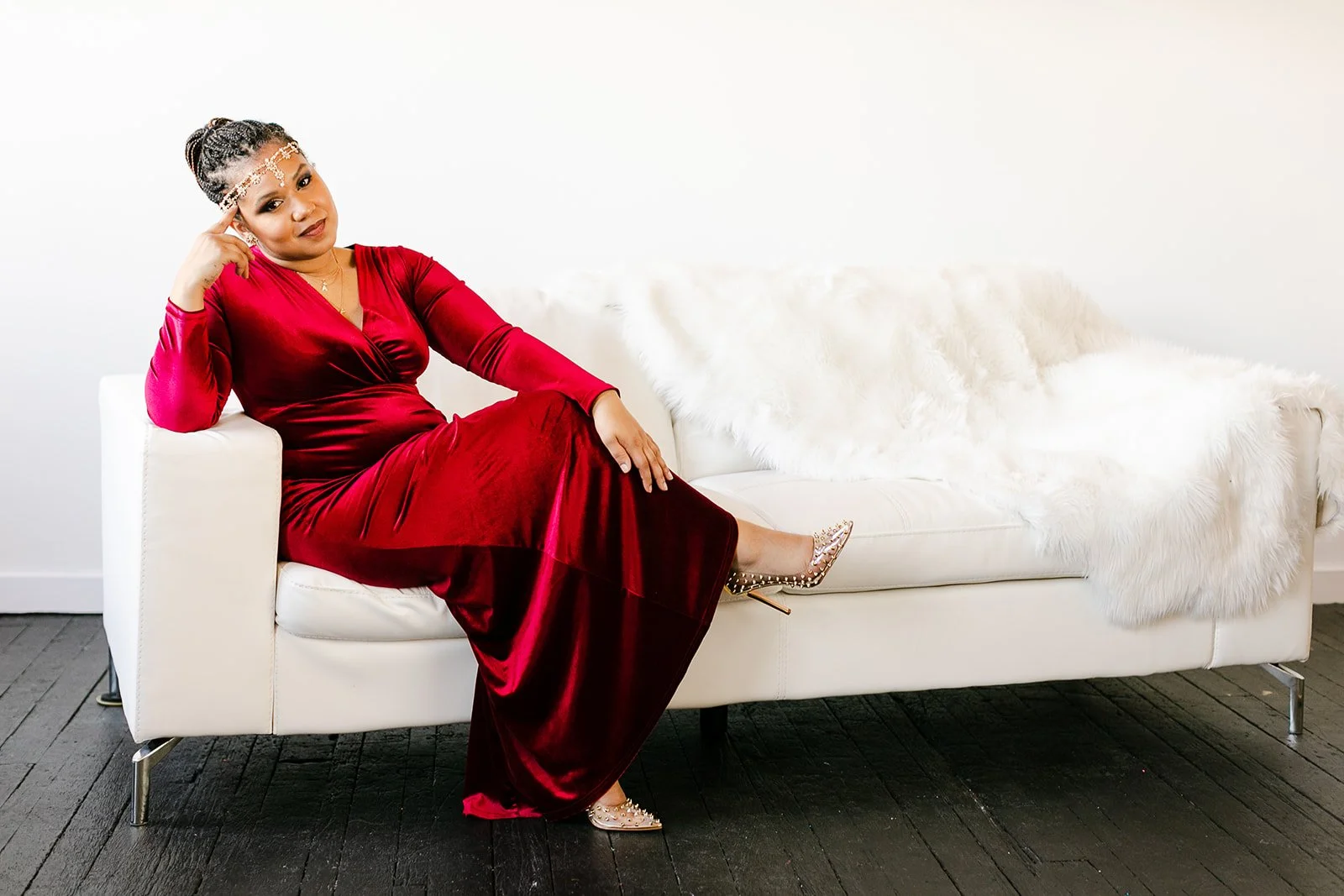 A beautiful black woman in a red velvet dress sitting on a white couch with a white furry blanket, wearing a beaded headband and gold studded high heels.