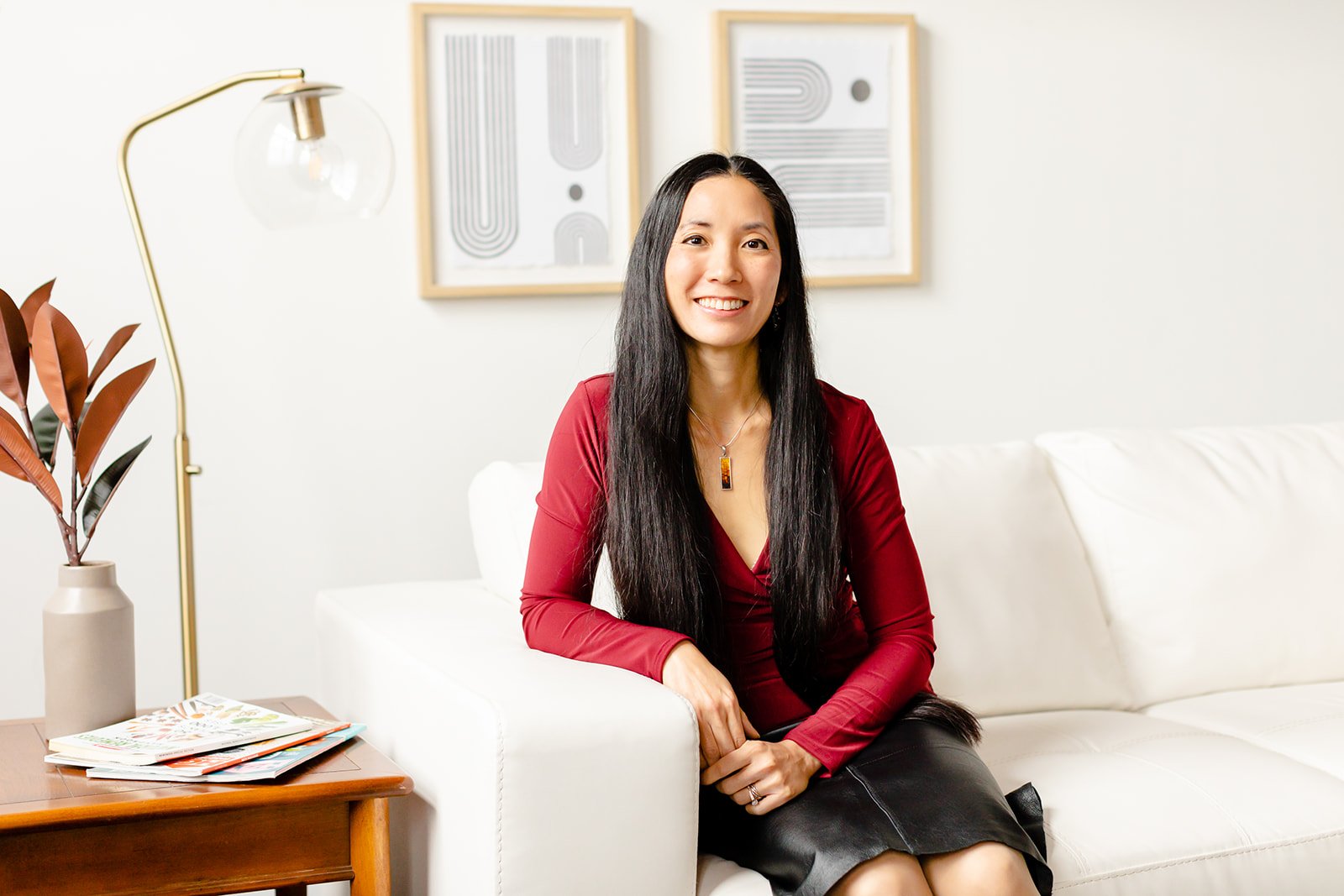 A beautiful Asian woman with long hair wearing a red shirt and a leather skirt poses on a white couch for a magazine feature