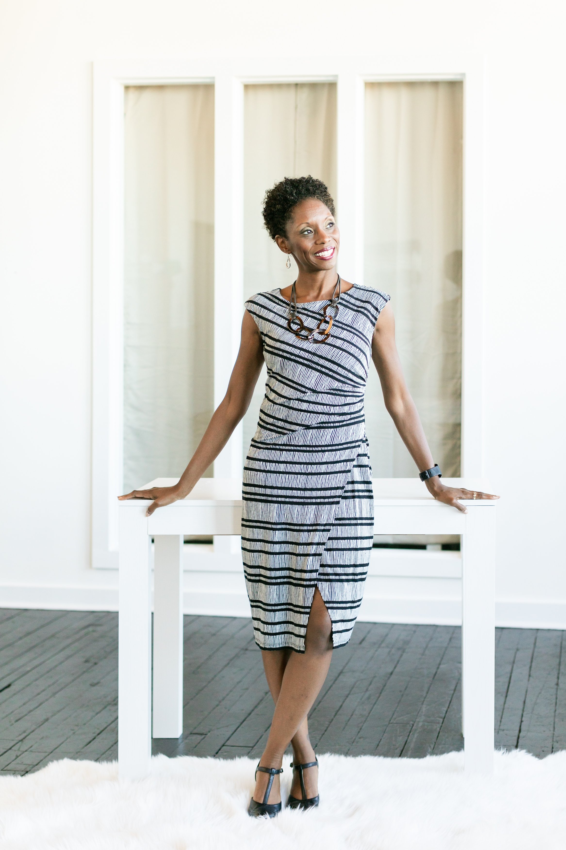 A black woman business owner stands proudly with her hands resting behind her on a white desk.