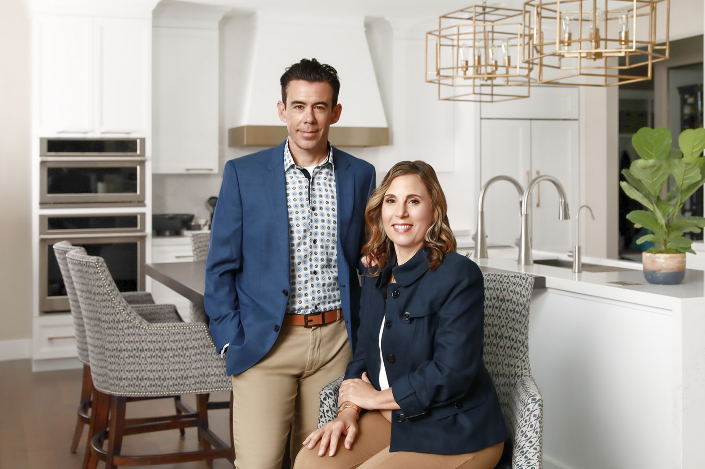 A husband-and-wife interior design and build team sit in a gorgeous white kitchen they remodeled.