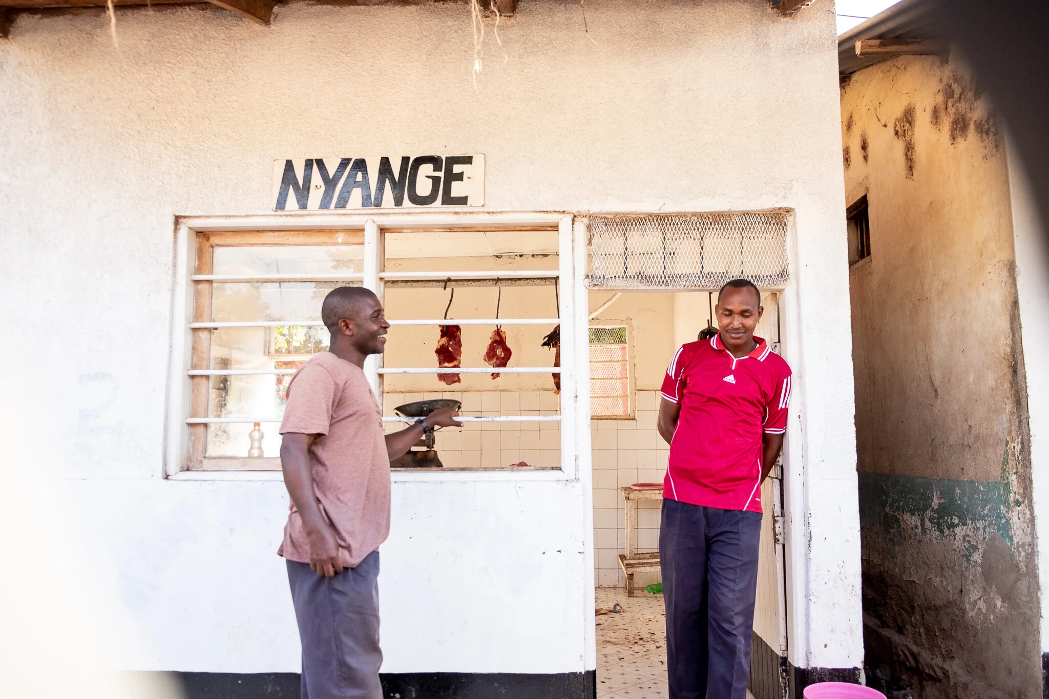 Two men standing outside a small meat shop with hanging meat in the background. One man, dressed casually in a brown t-shirt and gray trousers, is smiling and holding a plate, while the other man, dressed in a red sports shirt and dark trousers, stan