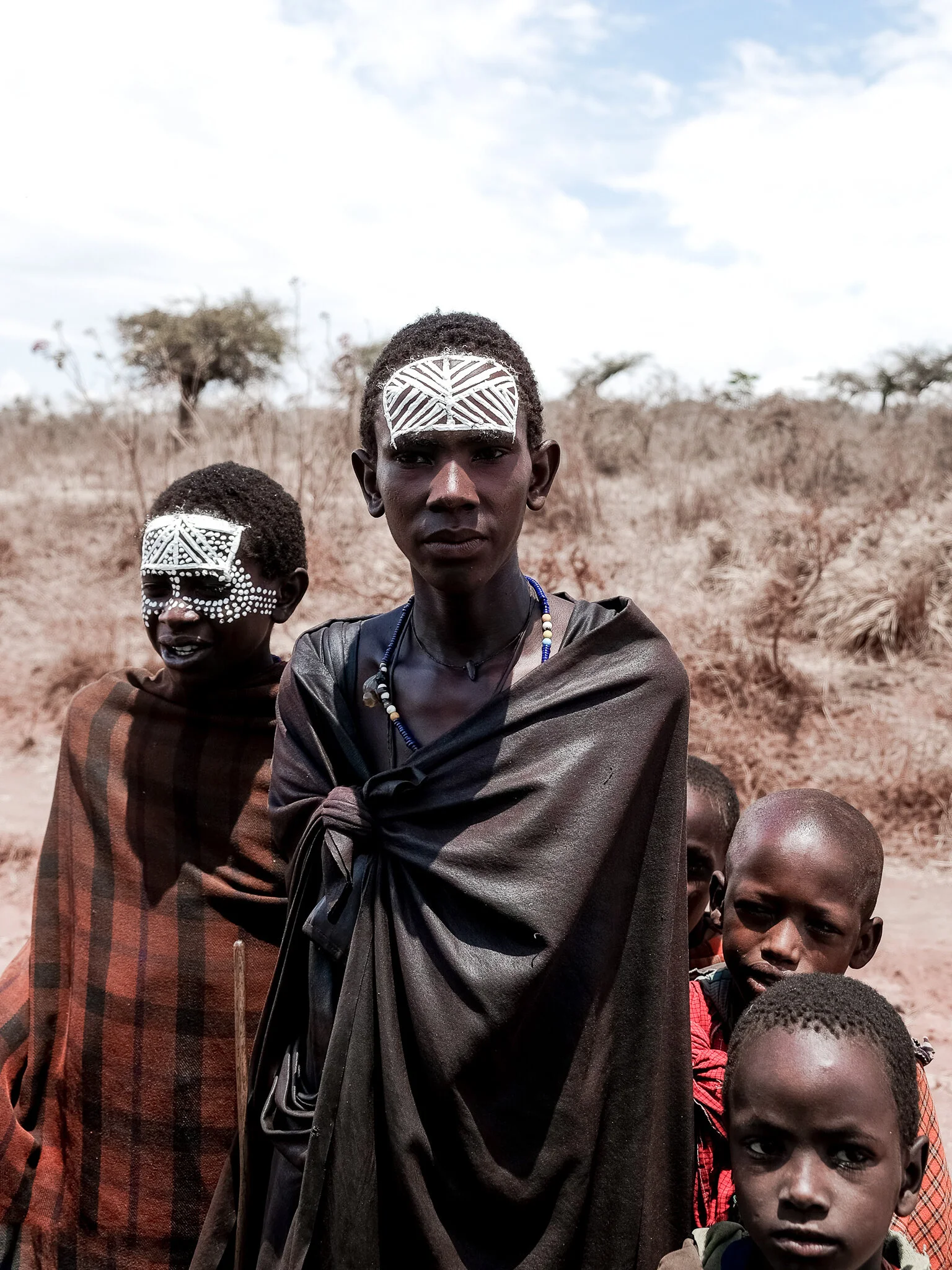 Group of Maasai children, some with traditional face paint, standing outdoors in a dry, sparse landscape with scattered trees.