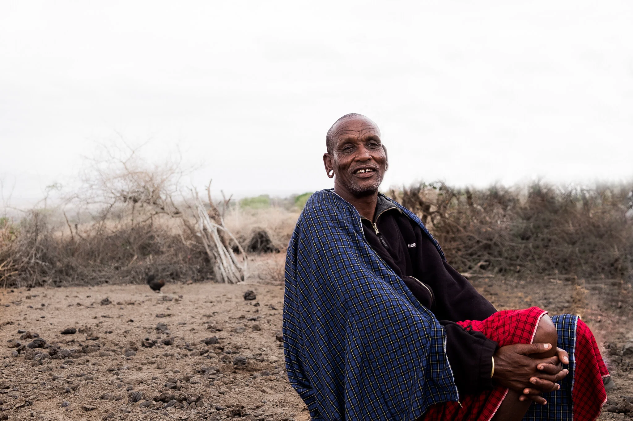 An elderly Maasai tribesman sitting on dry, rocky ground in a pastoral landscape with sparse bushes and leafless trees under a cloudy sky.