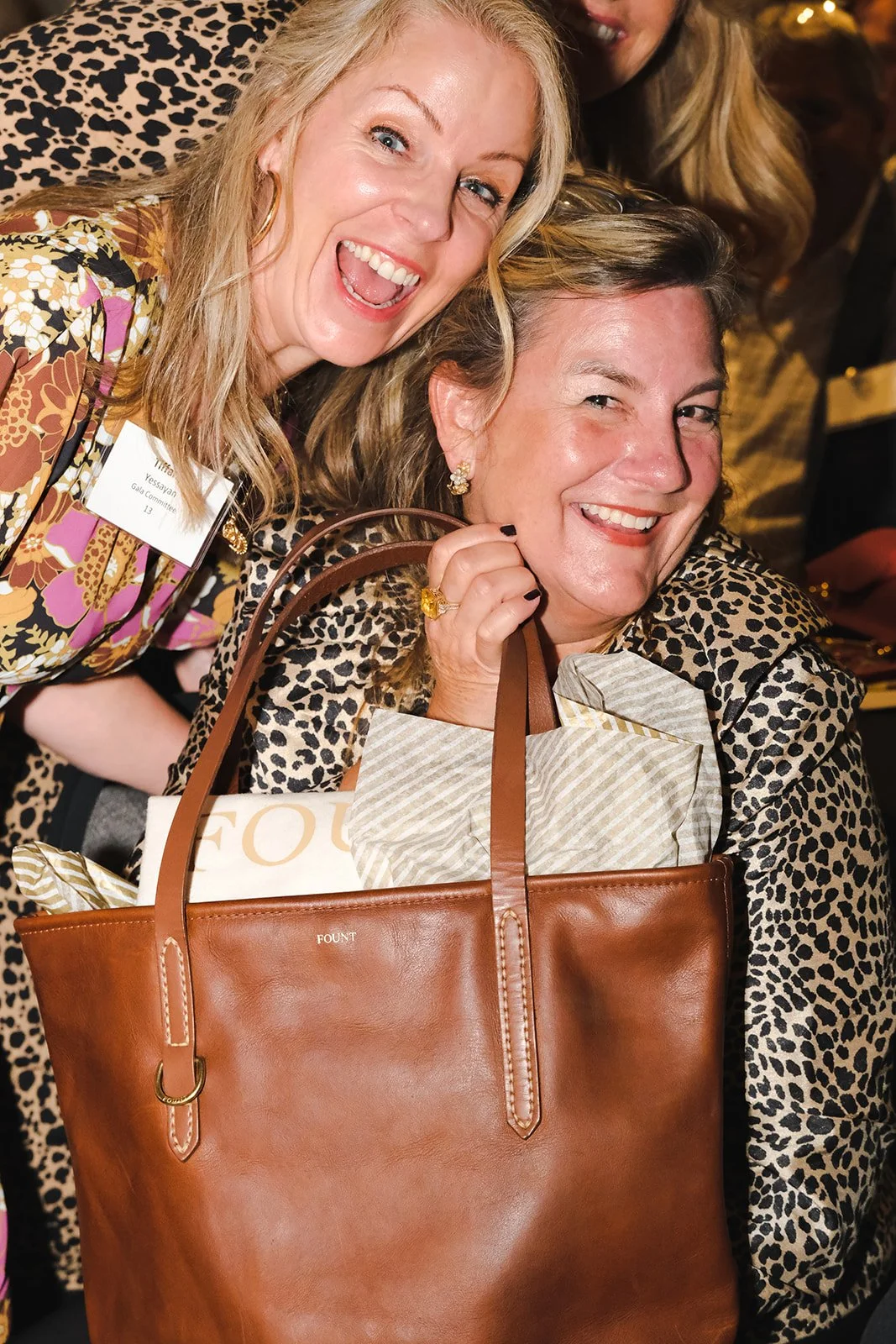 Two middle-aged women with blonde hair cozy up and smile at the camera after winning a FOUNT leather purse as the winning prize at a fundraising event.