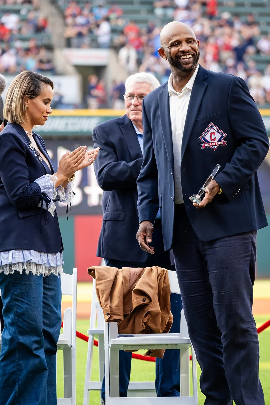 A man receiving an award at a sports stadium with two other people applauding him, one woman with her hands together and a man in glasses clapping.