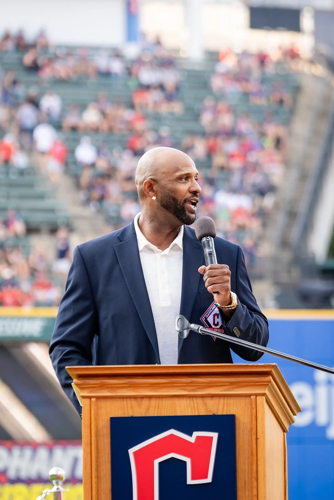 Hall of Fame Inductee CC Sabathia is speaking into a microphone at a podium with a Cleveland Guardians logo. He is dressed in a navy blazer and a white shirt, and is standing in a baseball stadium with blurred spectators in the background.