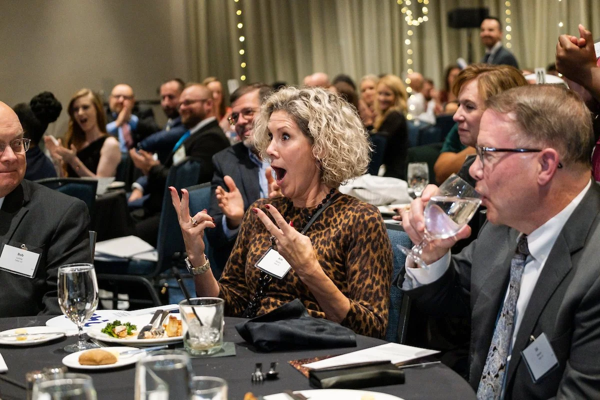 Woman with curly blonde hair in leopard print dress shows surprised expression at a formal event; surrounded by other attendees, some clapping, some drinking water, in a banquet hall with string lights.