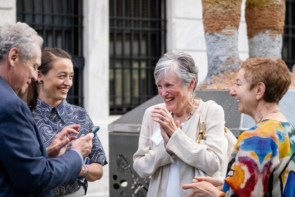 A woman with white hair surrounded by friends smiles and clasps her hands as she meets an artist for the first time at an event unveiling