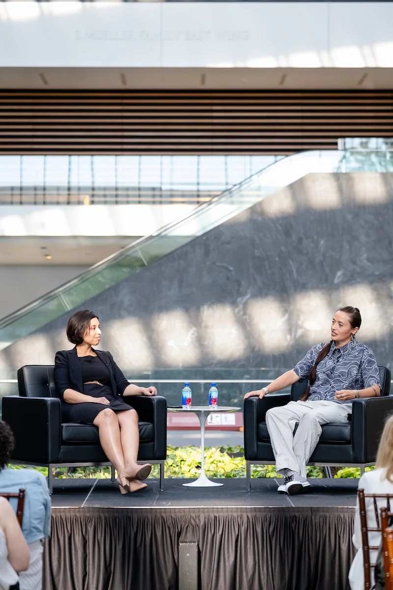 Two women are seated in black armchairs on a stage, engaging in a panelist conversation at an event. The background shows a modern, architecturally designed art museum with large windows and a glass railing.