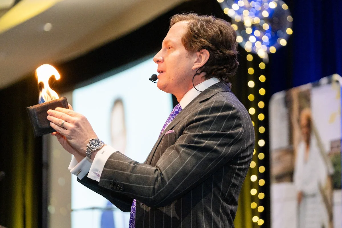 A man in a gray pinstripe suit holding a wallet with fire coming out of it during a presentation on stage.