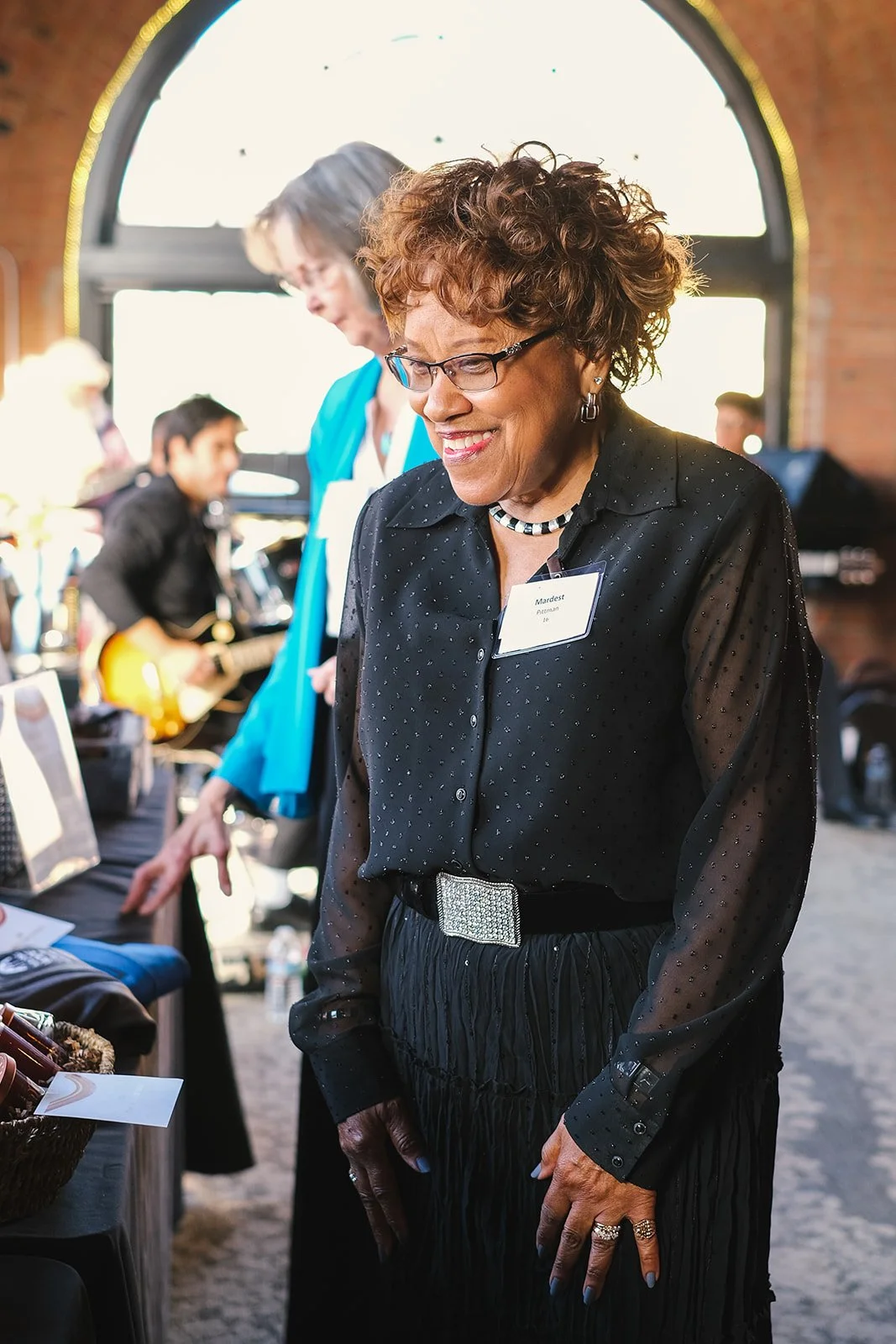 A smiling woman with curly hair, glasses, and jewelry stands at an indoor event, wearing a black blouse with polka dots and a large silver belt buckle. Behind her, people are interacting and playing guitar, with tables displaying items.