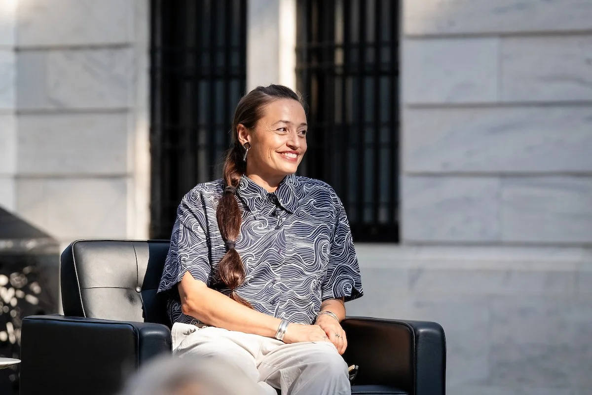Rose B. Simpson, a Mexican artist and sculptor sitting on a black chair outdoors, smiling, wearing a patterned shirt and light-colored pants, with a background of a stone building and windows at the Cleveland Museum of Art.