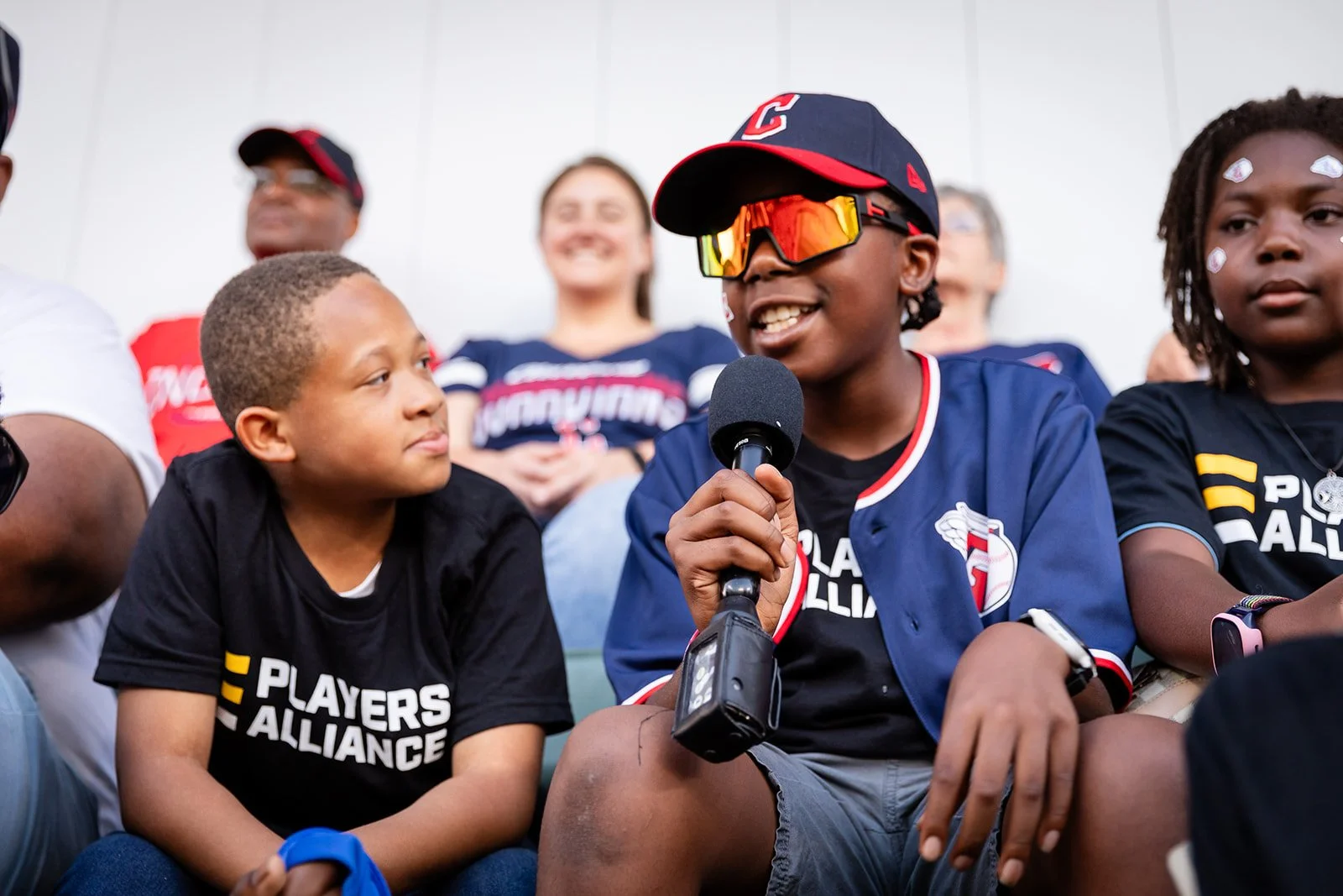 Children wearing Black Players Alliance T-shirts sitting on a row listening to a boy with sunglasses, cap, and jacket speaking into a microphone at a Cleveland Guardians baseball game.