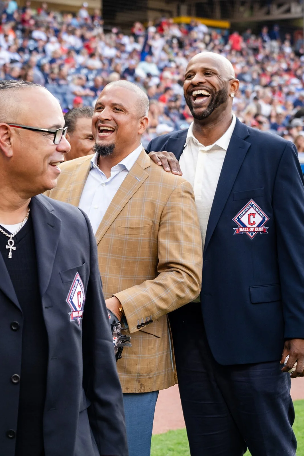 Three men standing, including CC Sabathia, and talking at a sports stadium, with a large crowd in the background. Two men are wearing jackets with a Cleveland Guardians Hall of Fame patch, and they are smiling and laughing.
