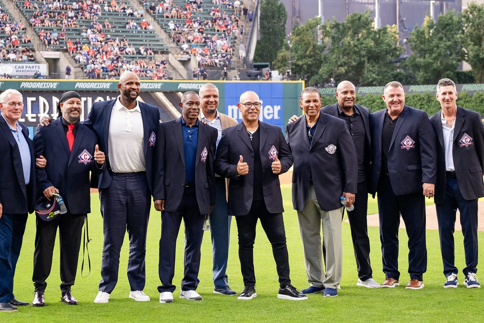 A group of Hall of Fame baseball players in suits standing on a baseball field, posing for a photo at the Cleveland Guardians stadium. They are smiling, some giving thumbs up. There are spectators in the stands behind them and a scoreboard above.