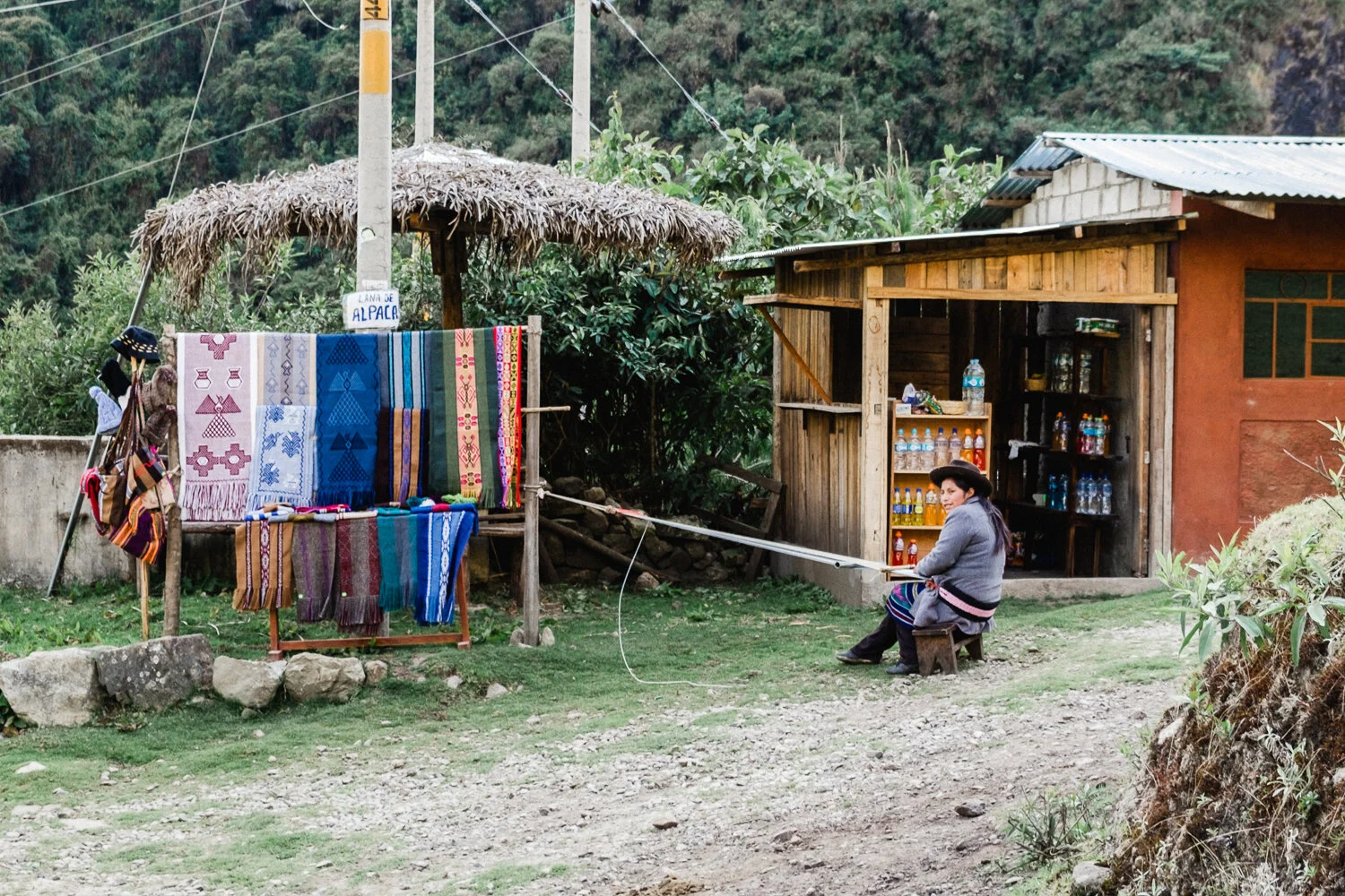 A woman sitting outside a small rustic store with bottled drinks, next to a stall displaying colorful woven textiles and traditional clothing, in a rural setting with green hills in the background.