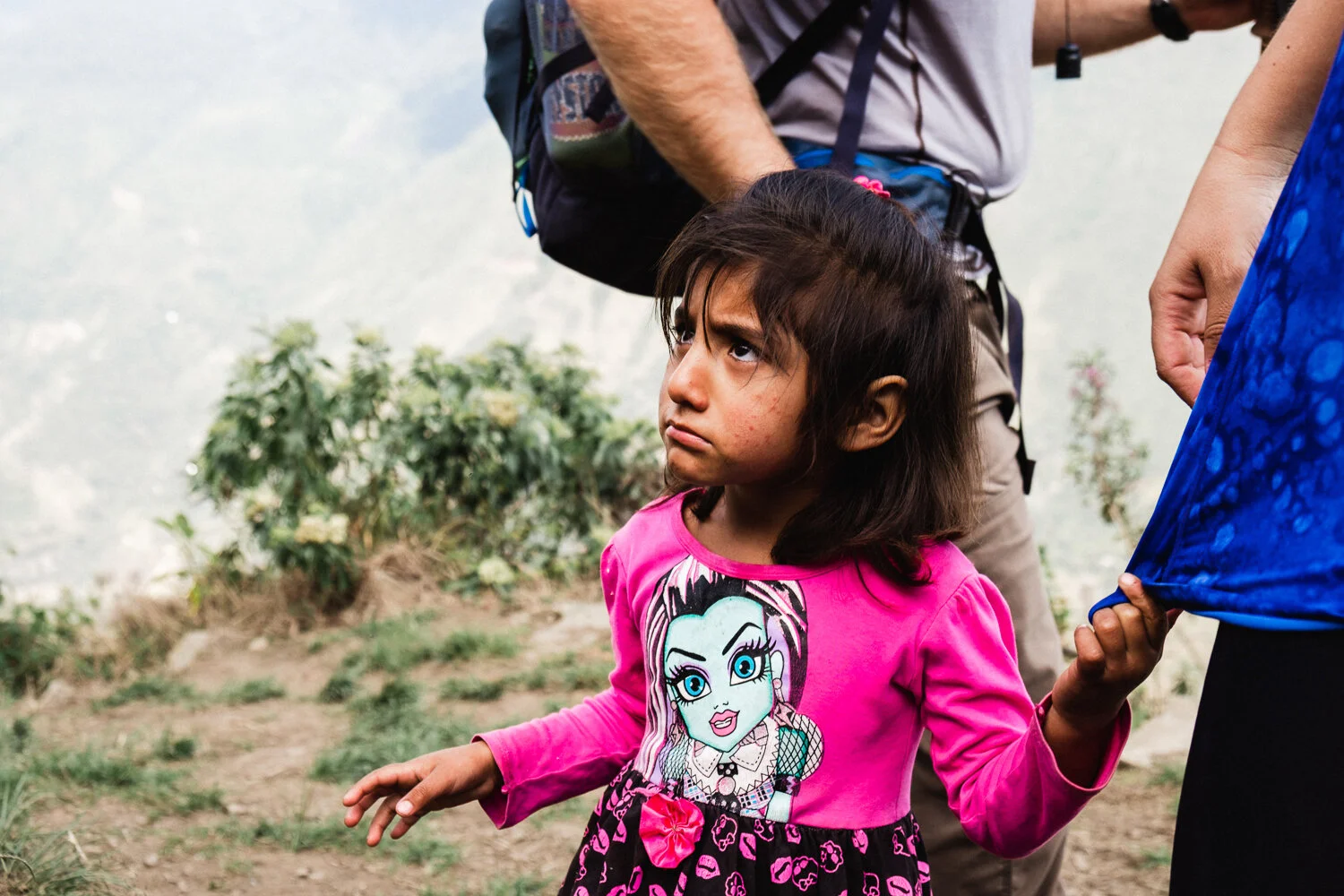A young girl with dark hair and a pink long-sleeve shirt featuring a cartoon character stands outdoors, looking upward with a serious expression. She is holding hands with an adult partially visible on the right, in a setting with distant mountains a