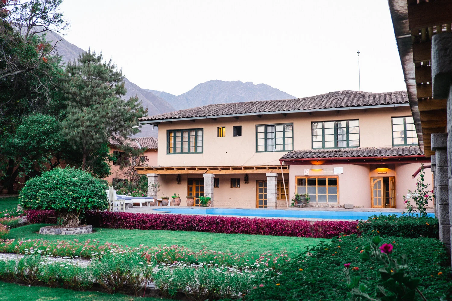 Backyard view of a two-story house with a tiled roof, large windows, and a swimming pool, surrounded by lush greenery, colorful flowers, and mountains in the background.