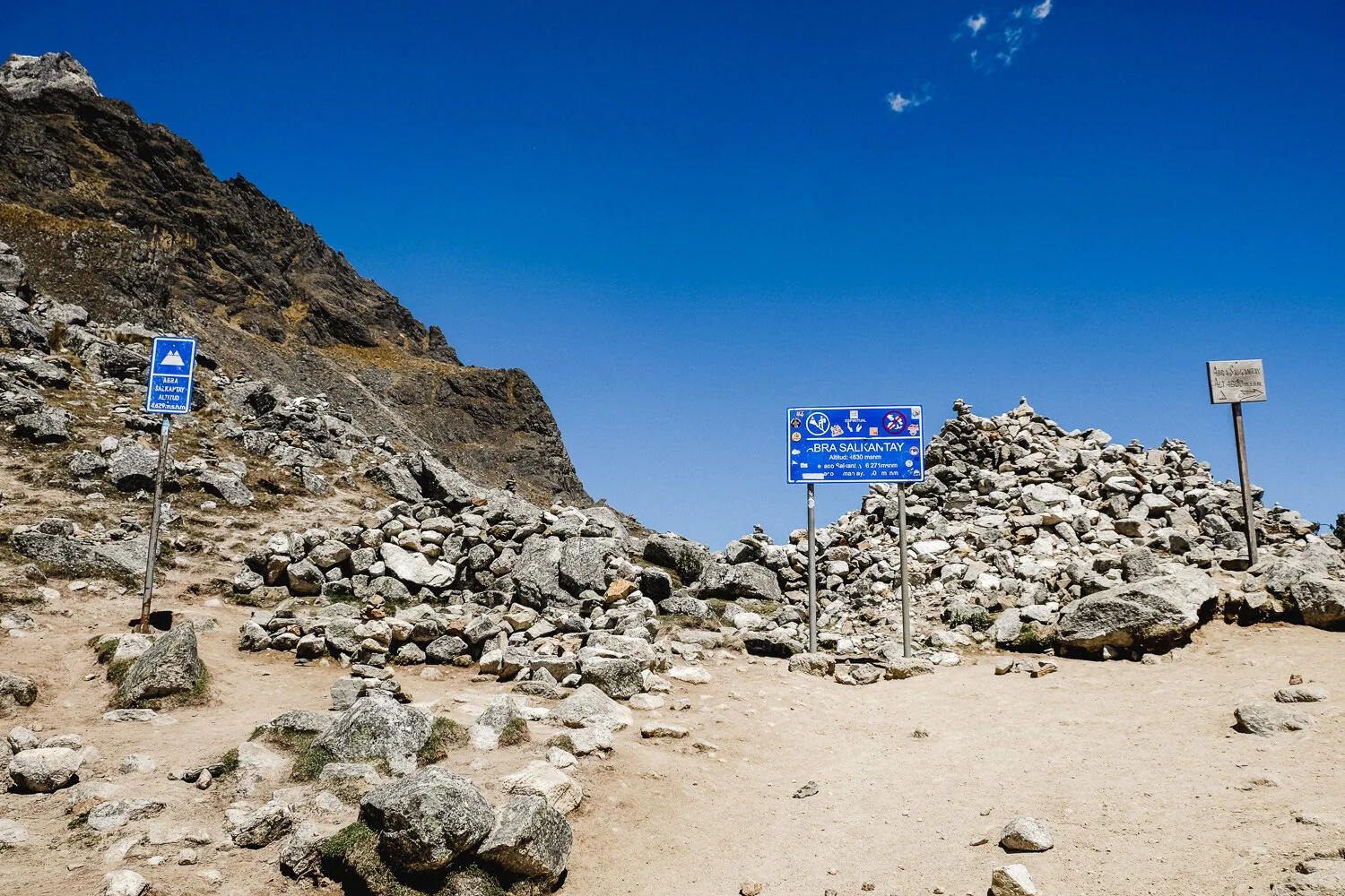 Mountain landscape with rocky terrain and blue sky, featuring multiple signs indicating the location is Abra Salikantay at 4,690 meters altitude.