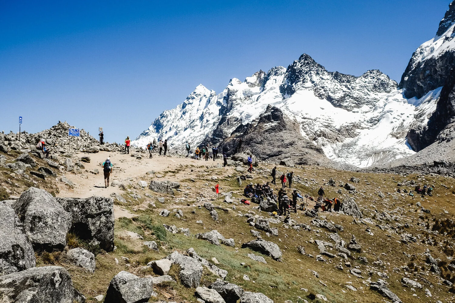 Group of hikers walking on a mountain trail with snow-covered peaks in the background.