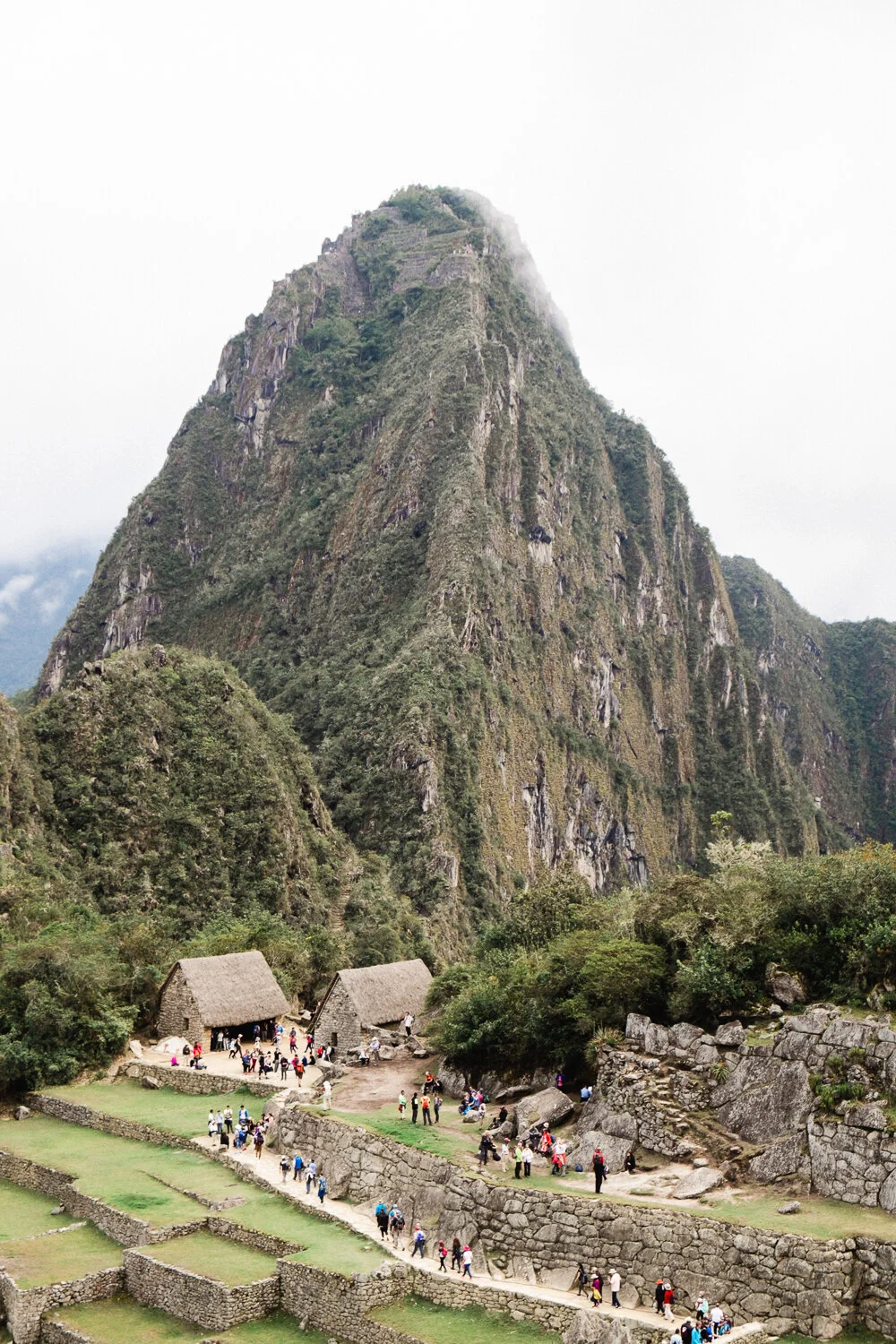 Tourists walking on stone pathways near small thatched-roof huts in Machu Picchu with foggy mountain in background.