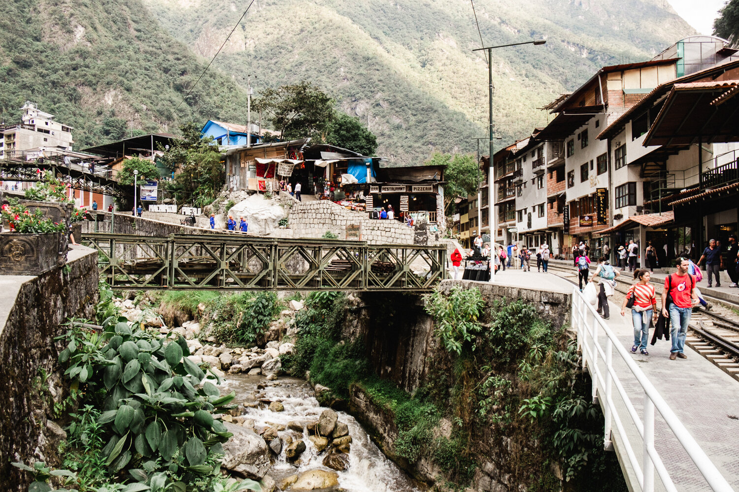 A busy street scene in a mountain town with people walking near shops and restaurants, a small bridge over a rocky stream, and hillside buildings with lush green mountains in the background.