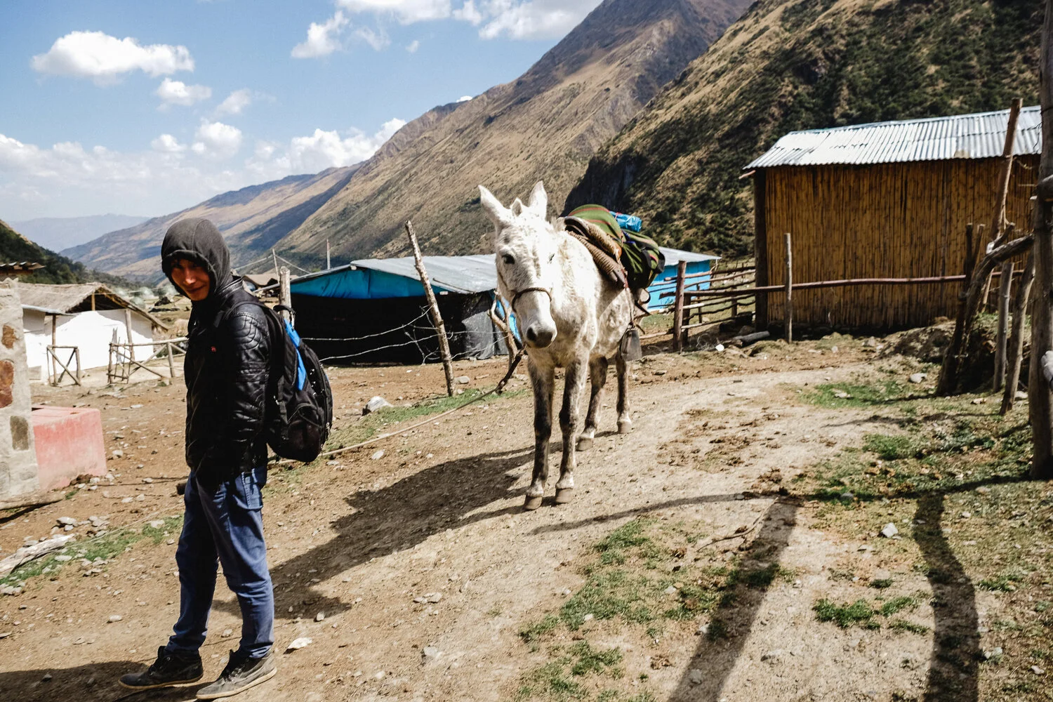 A person in a black jacket and blue pants smiling with a backpack, standing next to a donkey with a loaded pack, in a mountainous rural area with small houses and fences.