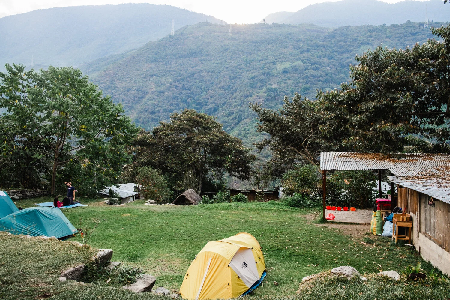 A campsite with tents set up on a grassy area surrounded by trees and mountains in the background, with people sitting near the trees.