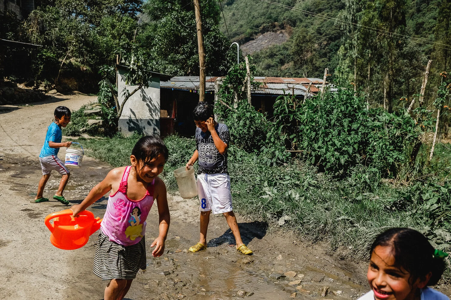 Children playing and washing outdoors on a dirt road in a rural area, with a small house and greenery in the background.