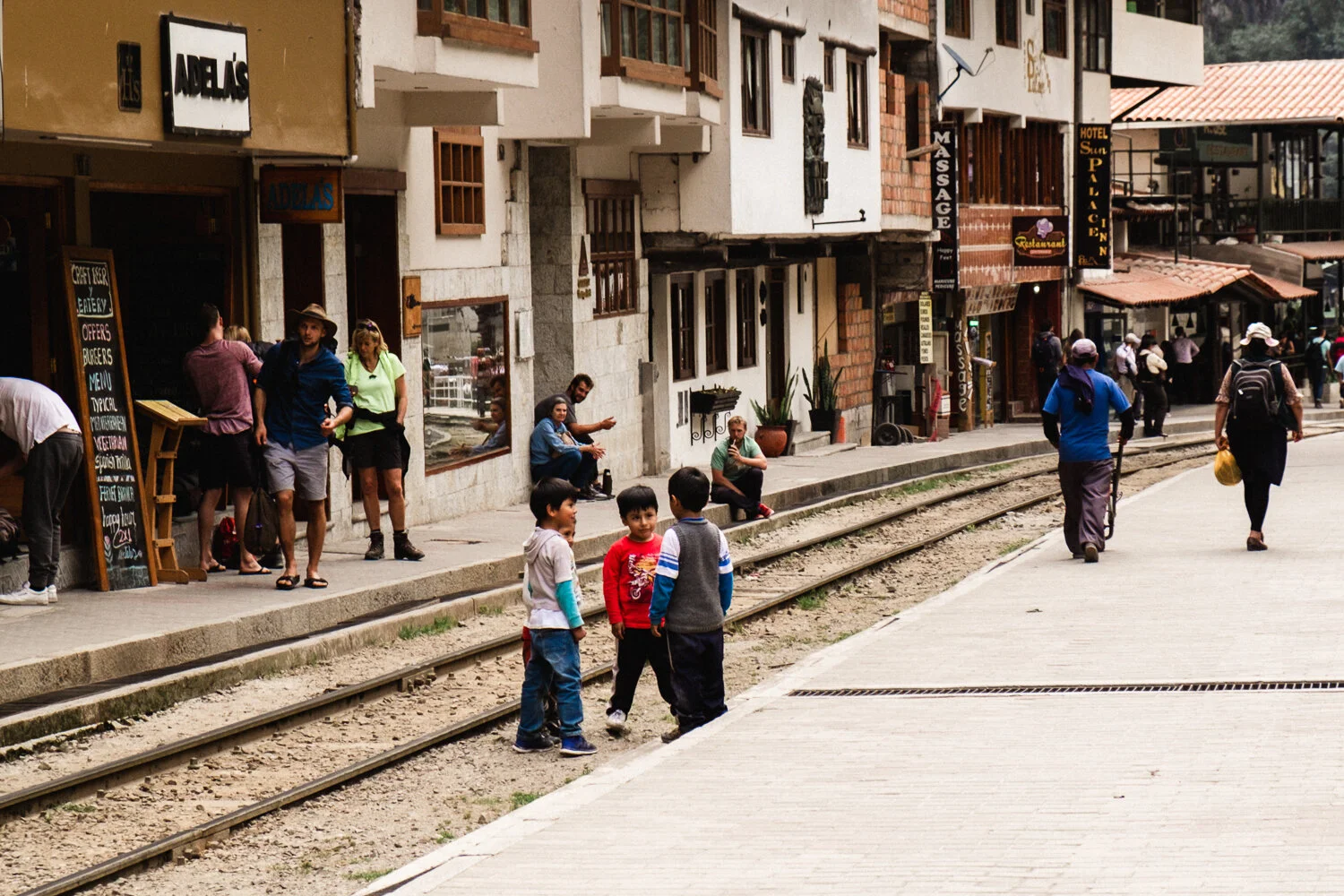 Street scene with children playing near train tracks, pedestrians walking and sitting outside shops, buildings with signs for massage, hotel, and restaurant, people carrying bags, and a tram in the background.