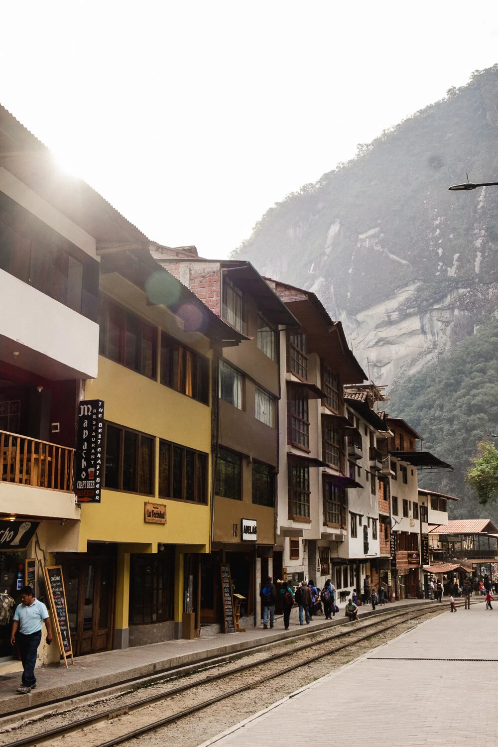 Colorful buildings along a street with tram tracks, and a mountainous landscape in the background of Aguas Calientes.