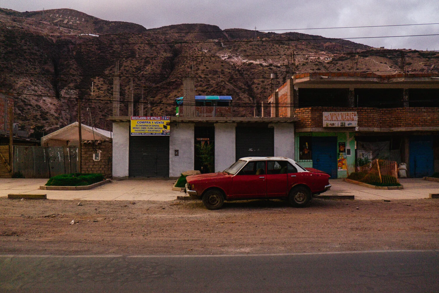 A street scene with a vintage red car parked in front of small shops and houses, with mountains in the background under a cloudy sky.