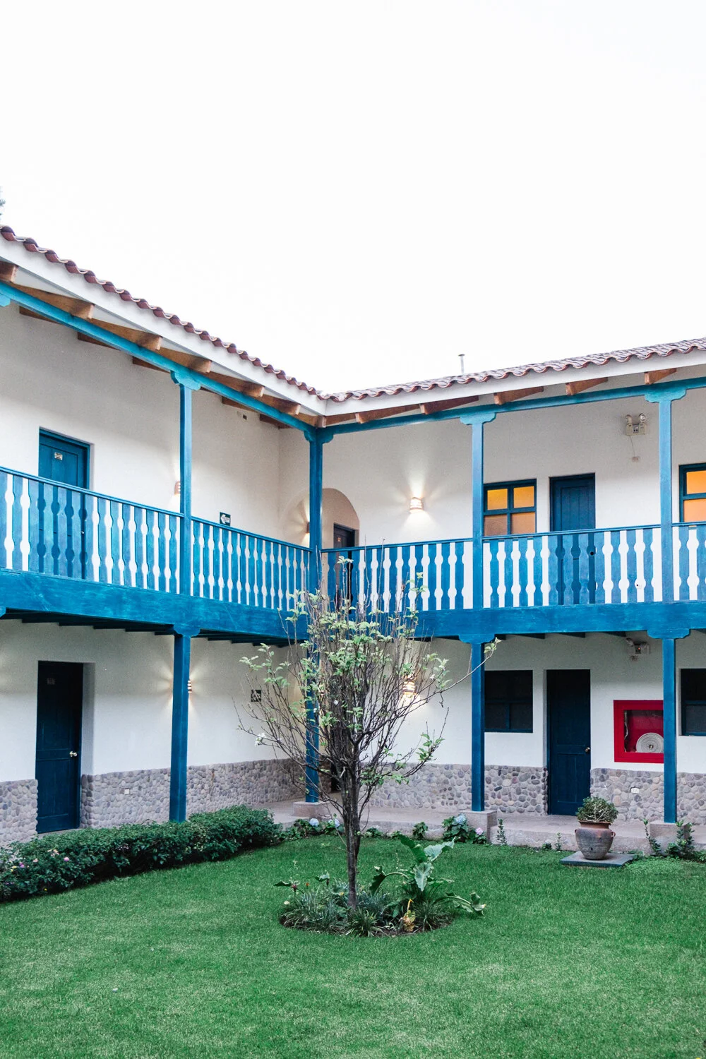 Two-story courtyard with white walls, blue wooden balconies and doors, a small tree in the center, and a manicured lawn.