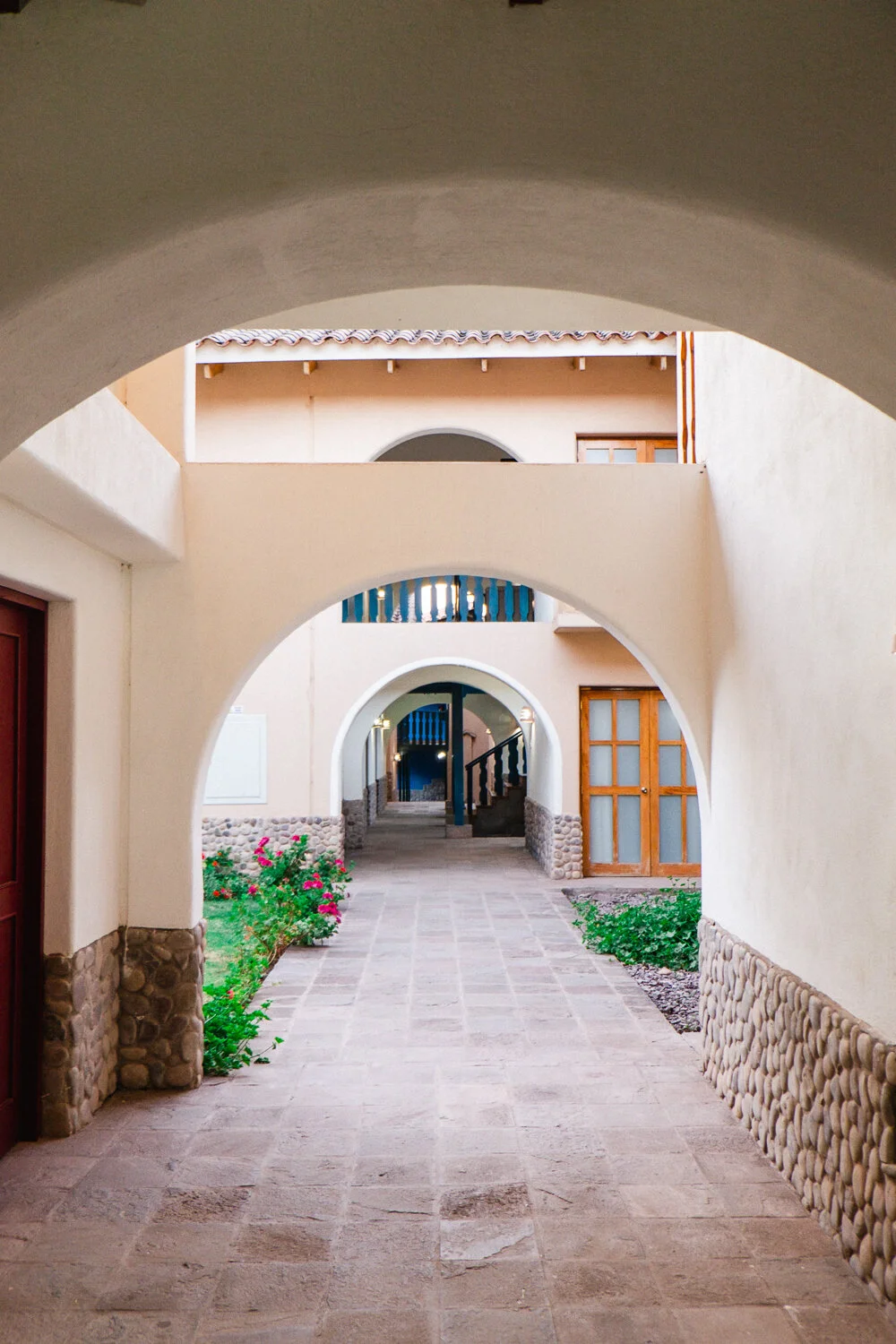 Archways leading to a courtyard with stone-paved walkway, beige walls, wooden doors, and flower beds with pink flowers.