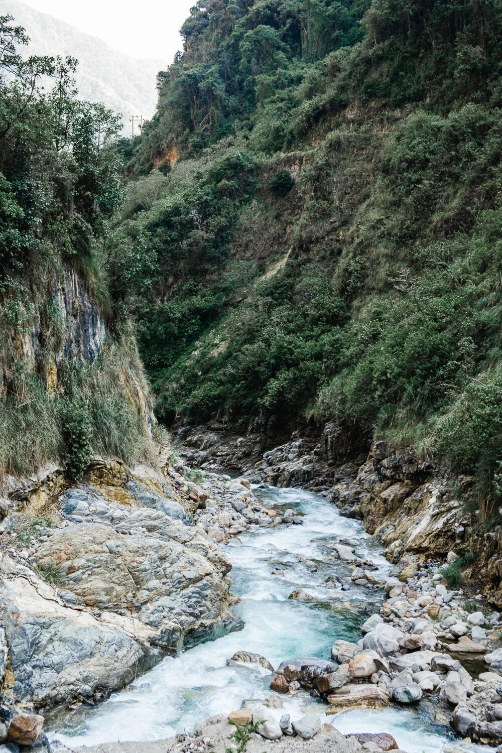 A narrow mountain river flowing through a lush green canyon with steep rocky slopes.