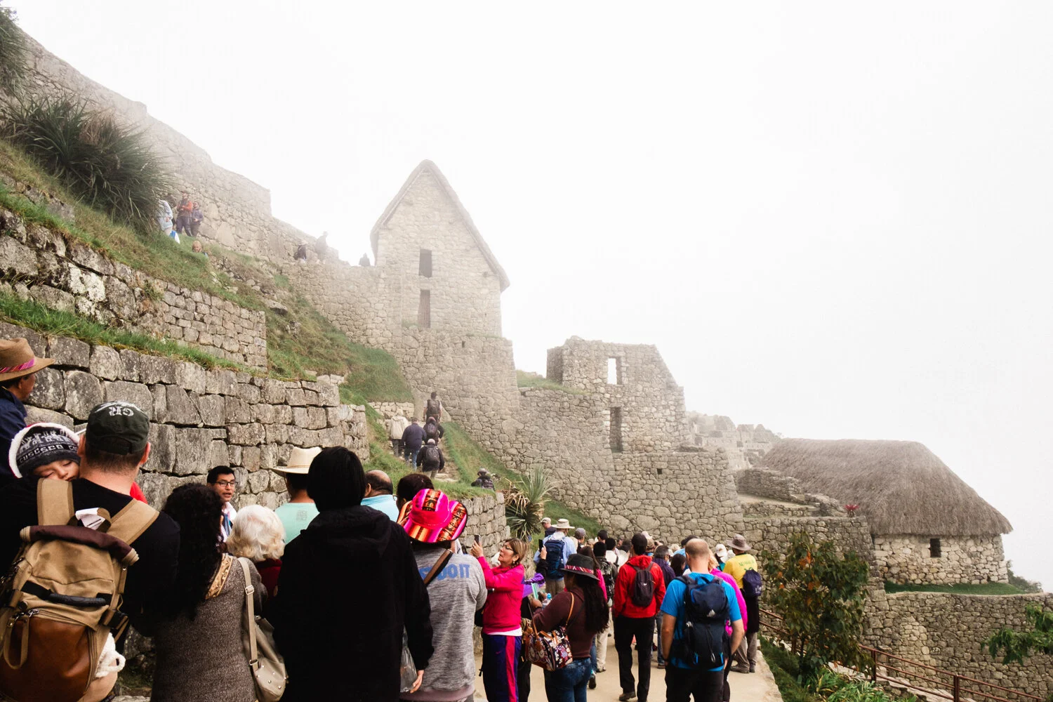 Tourists explore Machu Picchu with ancient stone structures and terraced landscape in foggy weather.
