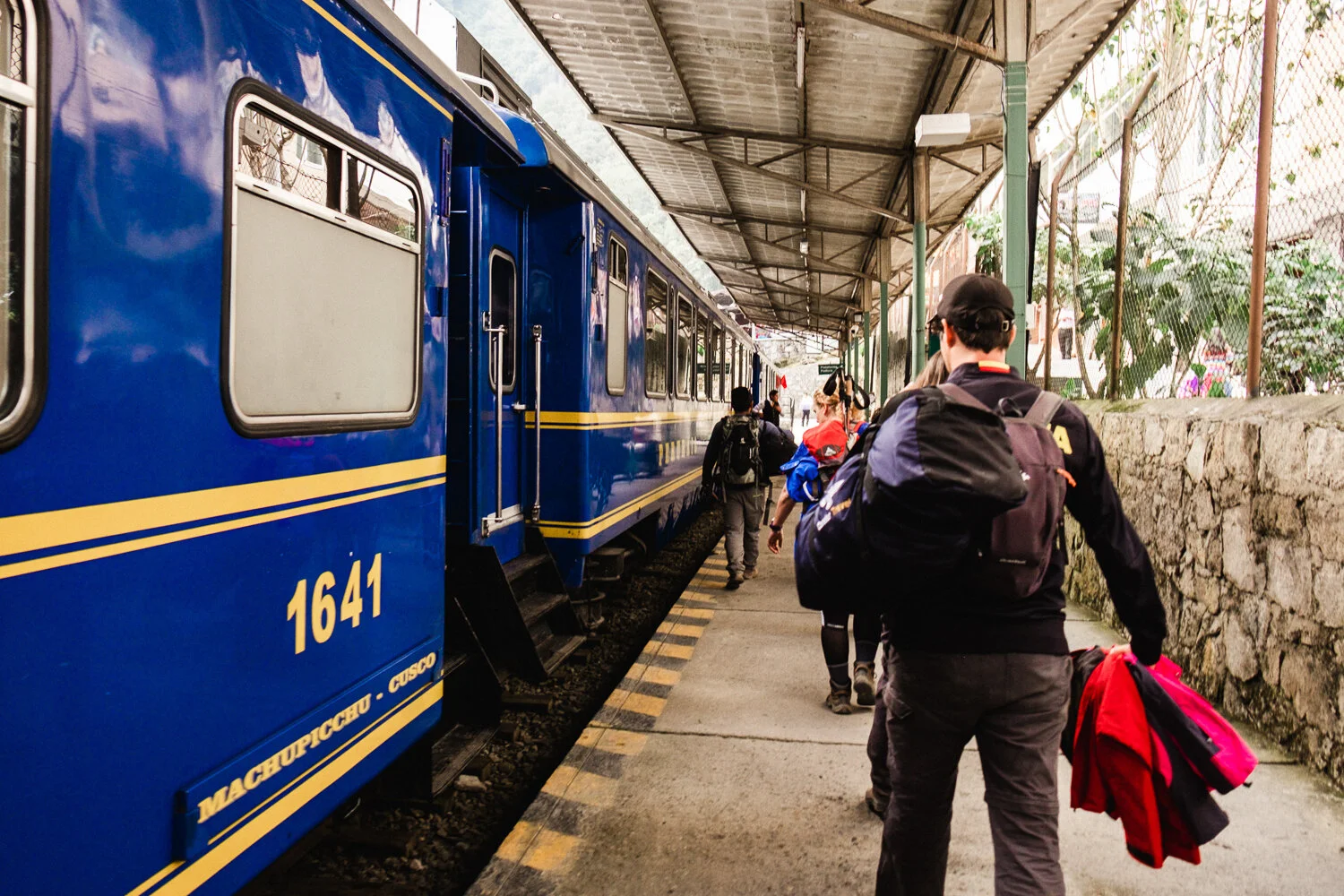 People boarding a blue train at a station platform in Aguas Calientes, Peru, with a stone wall and fencing on the side.
