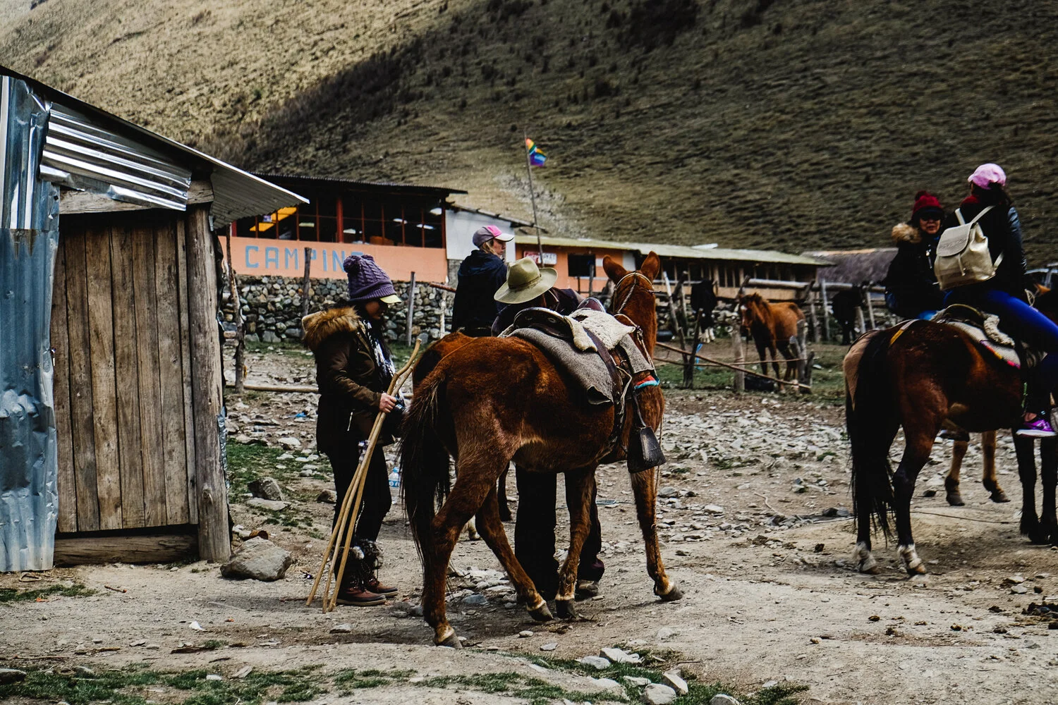 Group of people with horses at a mountain camp, some riding and some standing next to horses, with a rustic wooden building and hillside in the background.